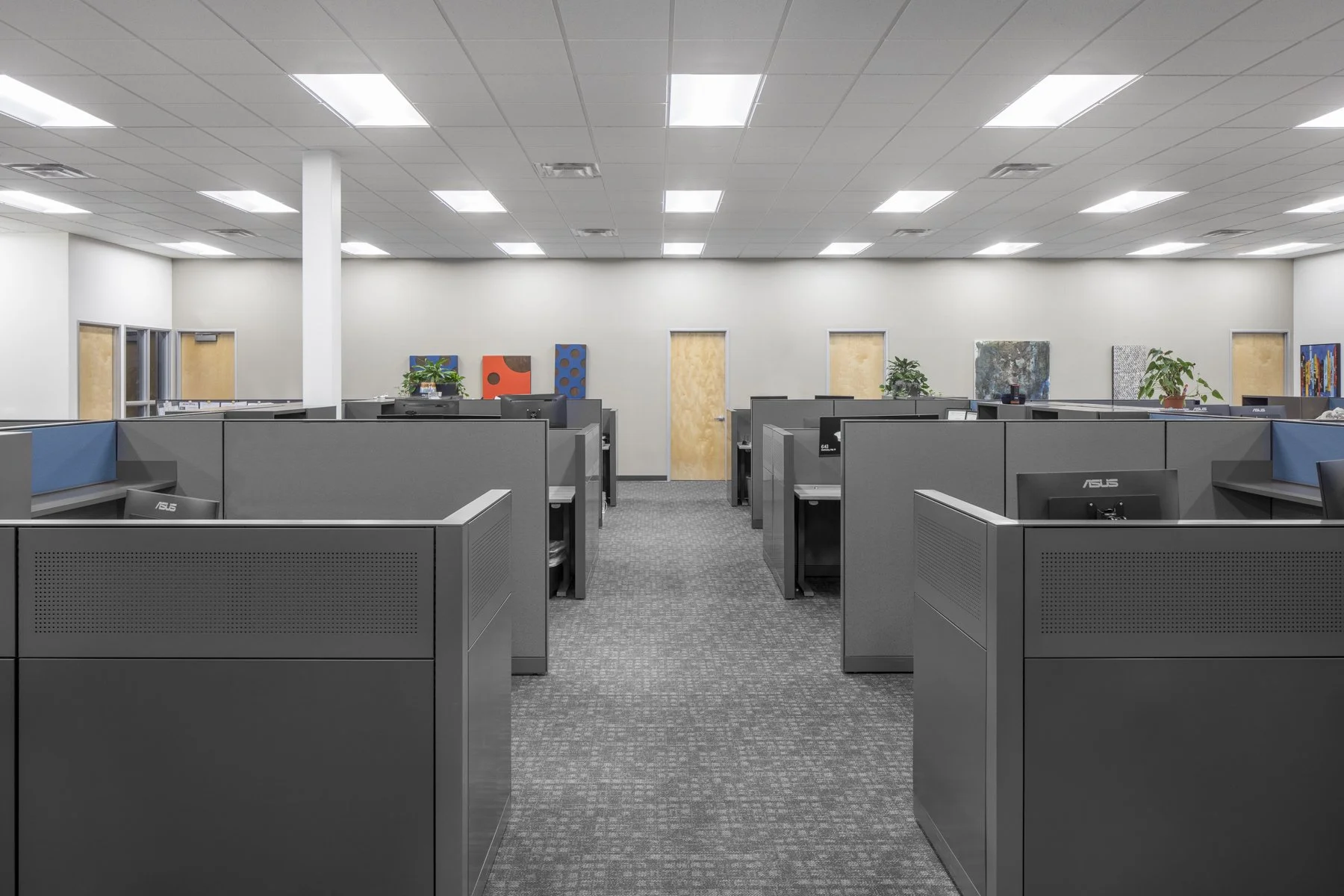 Empty office cubicles with desktop computers, plants, and colorful artwork on the wall, under bright ceiling lights.