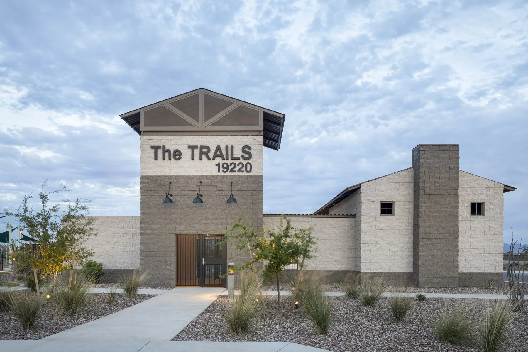 Exterior view of a modern building with a sign reading 'The TRAILS 19220,' featuring neutral-colored stone walls, a small landscaped yard with desert plants, and a cloudy sky in the background.