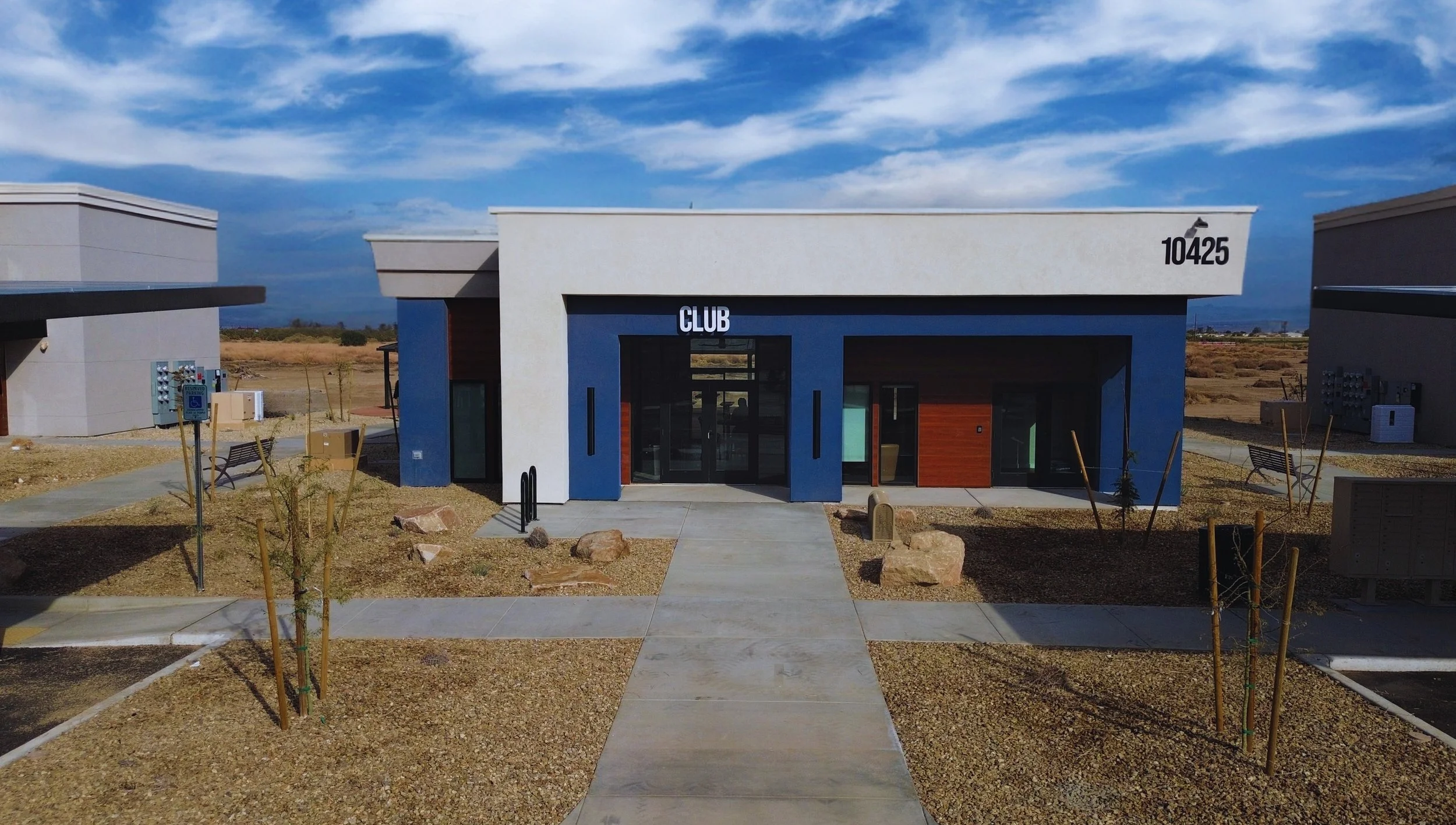 Modern building with blue and white facade, labeled 'CLUB' and number '10425', with a pathway leading to entrance, surrounded by dry landscape and benches under a partly cloudy sky.