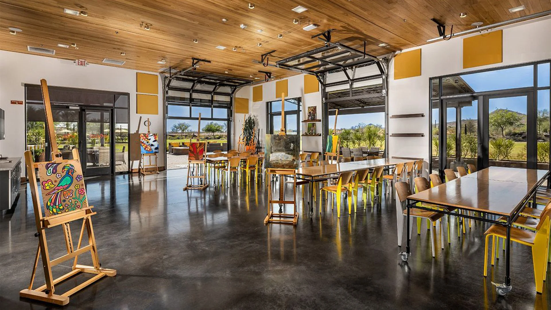 An art studio with easels, paintings, tables, and chairs, overlooking a scenic outdoor landscape through large windows.