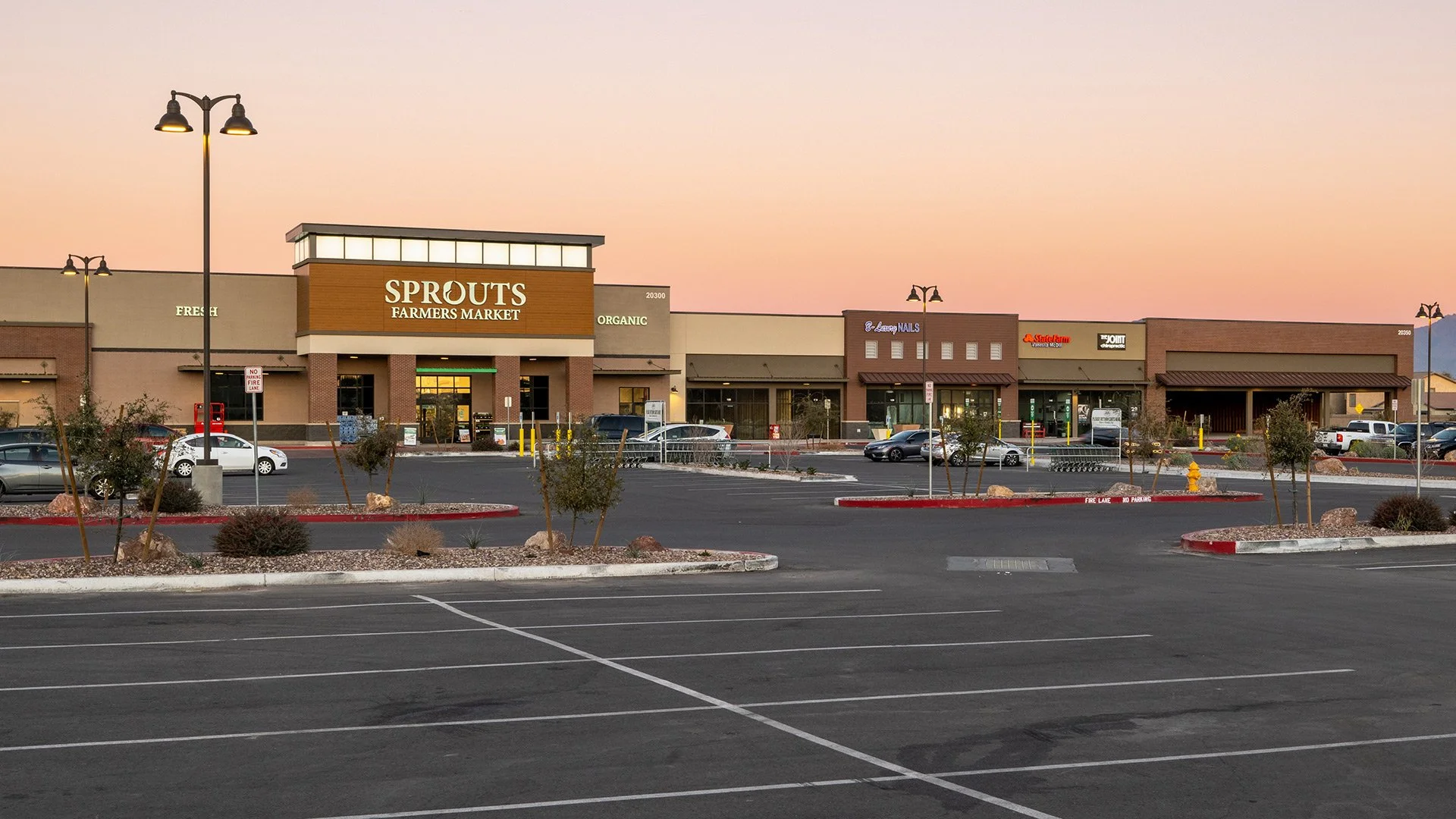 A strip mall parking lot at sunset with storefronts including Sprouts Farmers Market, nail salon, and other retailers, with a mostly empty parking lot and small landscaped islands.