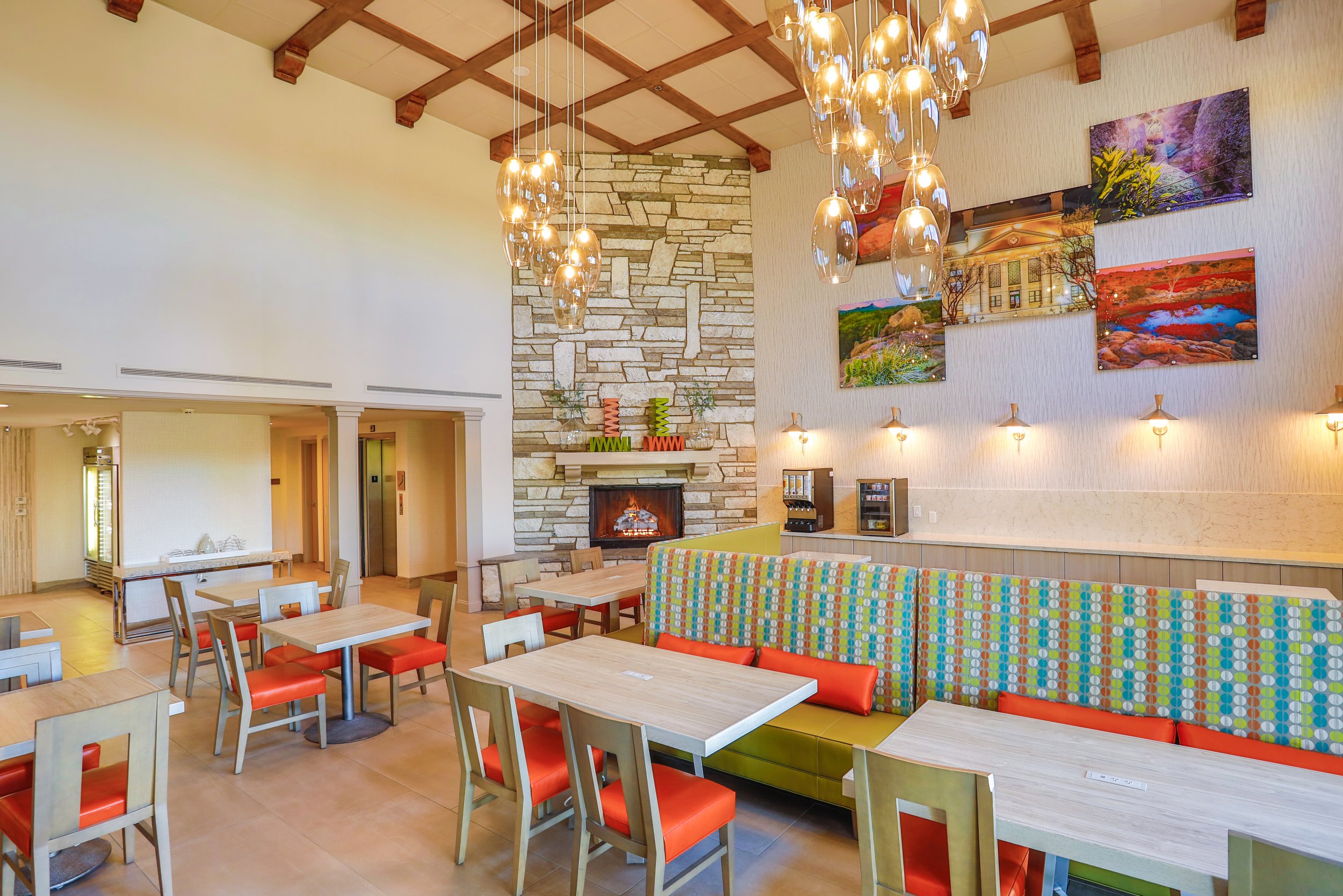 A spacious, brightly lit cafe with colorful decor, a stone fireplace, wall art, and a seating area with tables, chairs, and a long booth with patterned fabric.