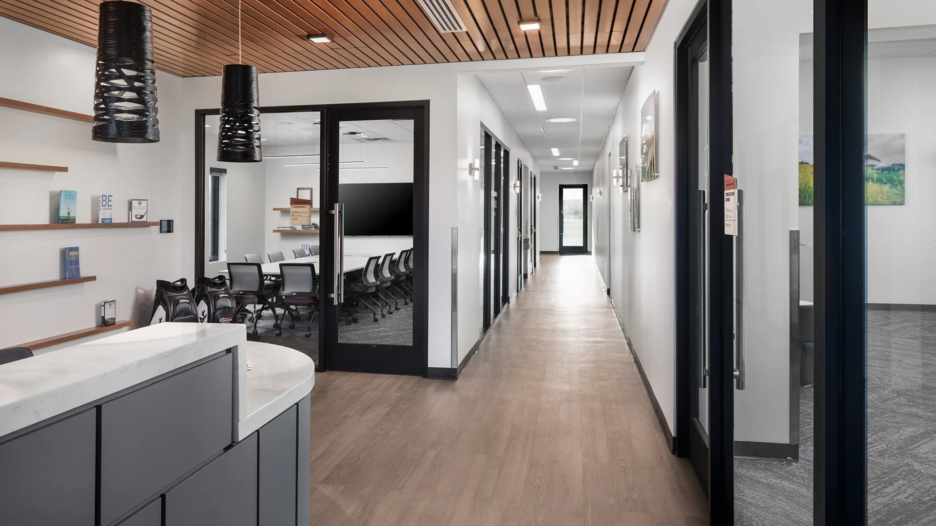 Modern conference room with a long table and chairs, overlooking a hallway with framed pictures, and a reception area with bookshelves and hanging pendant lights.