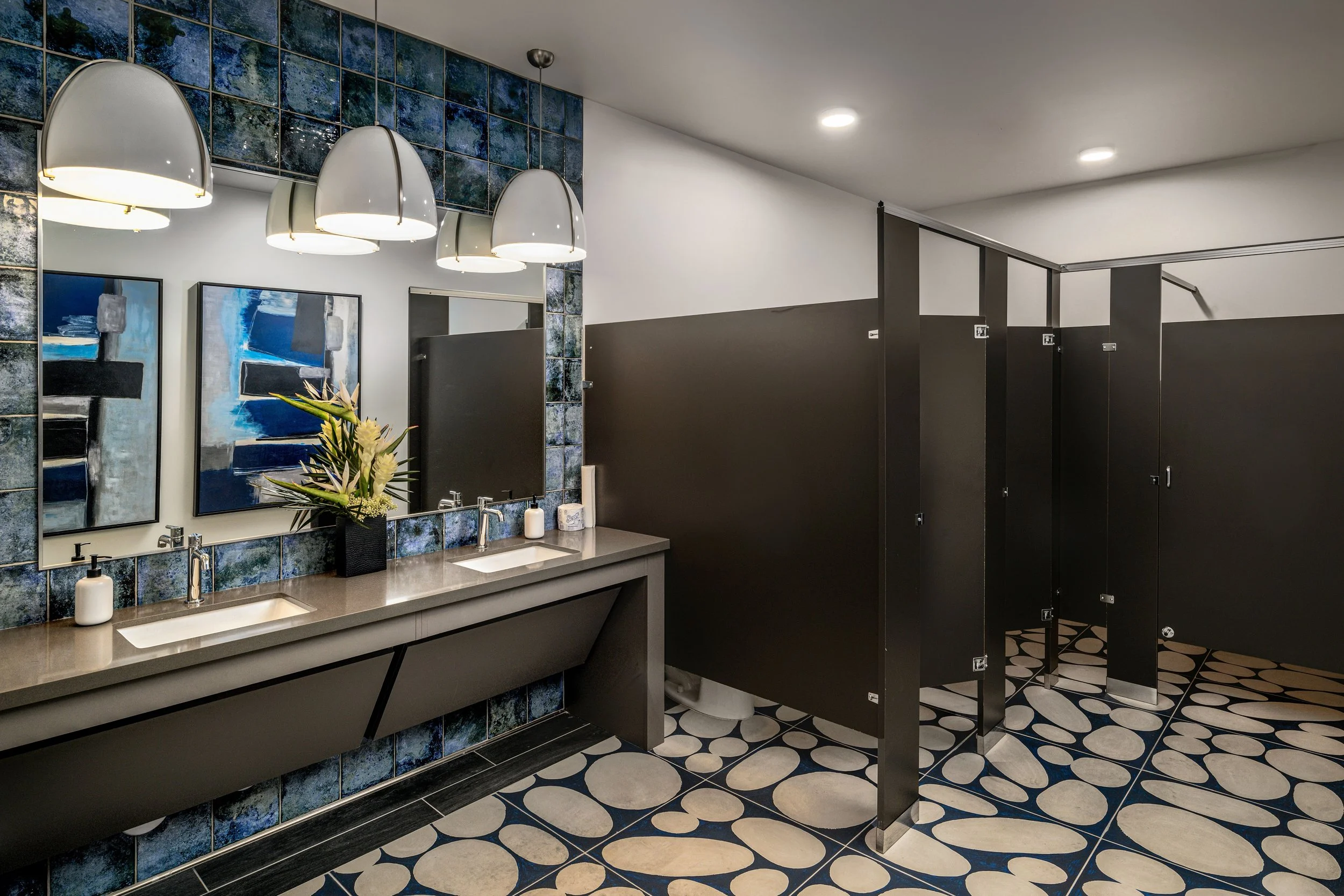 Public women's bathroom with two sinks, large mirror, modern lighting, black and blue tile decor, and three black stalls.