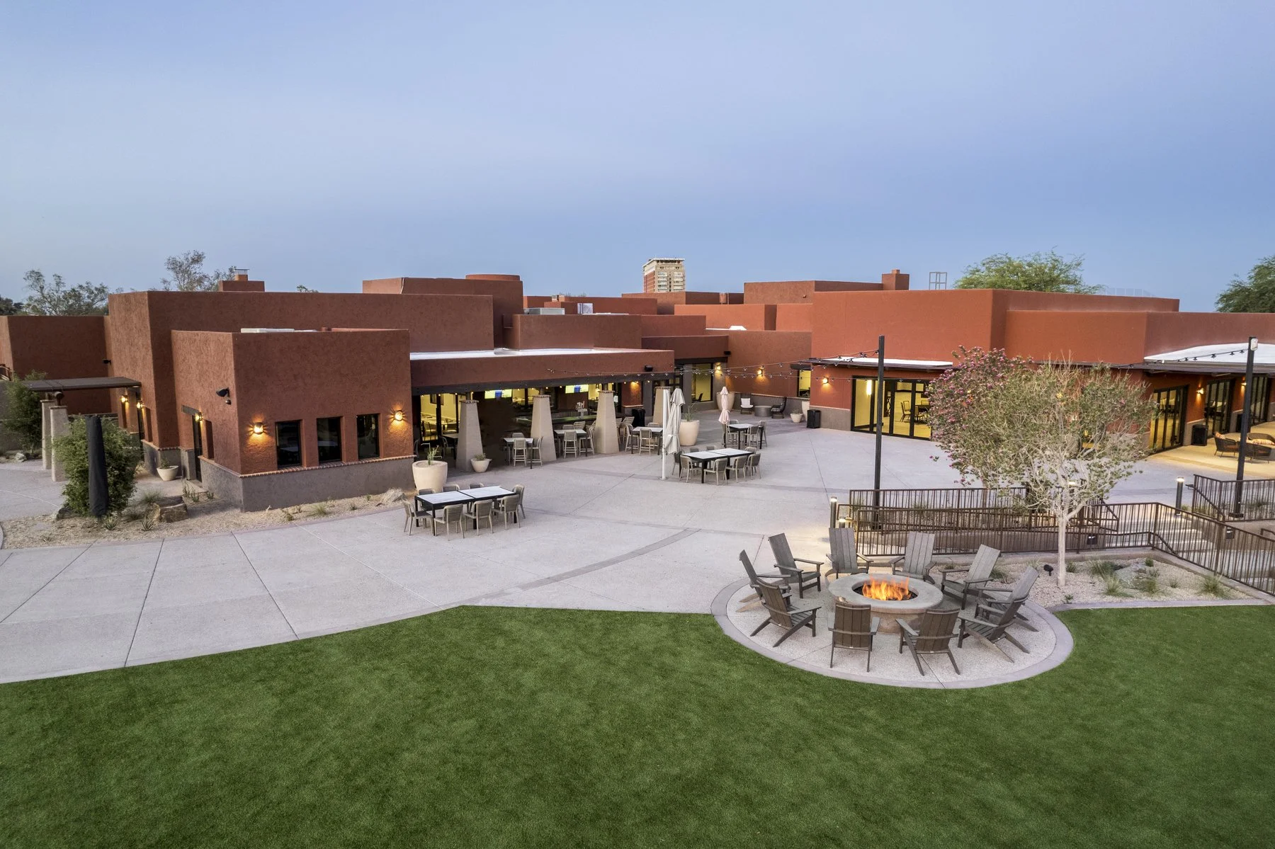Outdoor patio with tables, chairs, umbrellas, a fire pit, and a tree, in front of a modern building with a red exterior and large glass windows, during dusk.