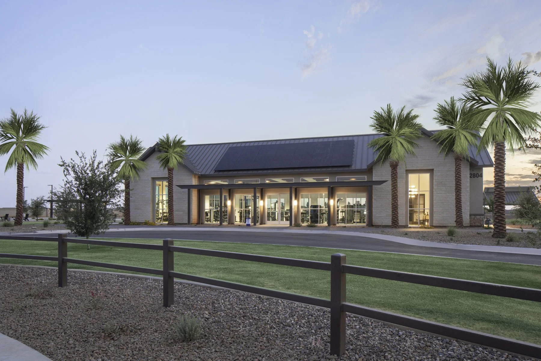 Modern building with solar panels on roof, large glass windows, surrounded by palm trees, with a green lawn and a wooden fence in the foreground, during dusk.