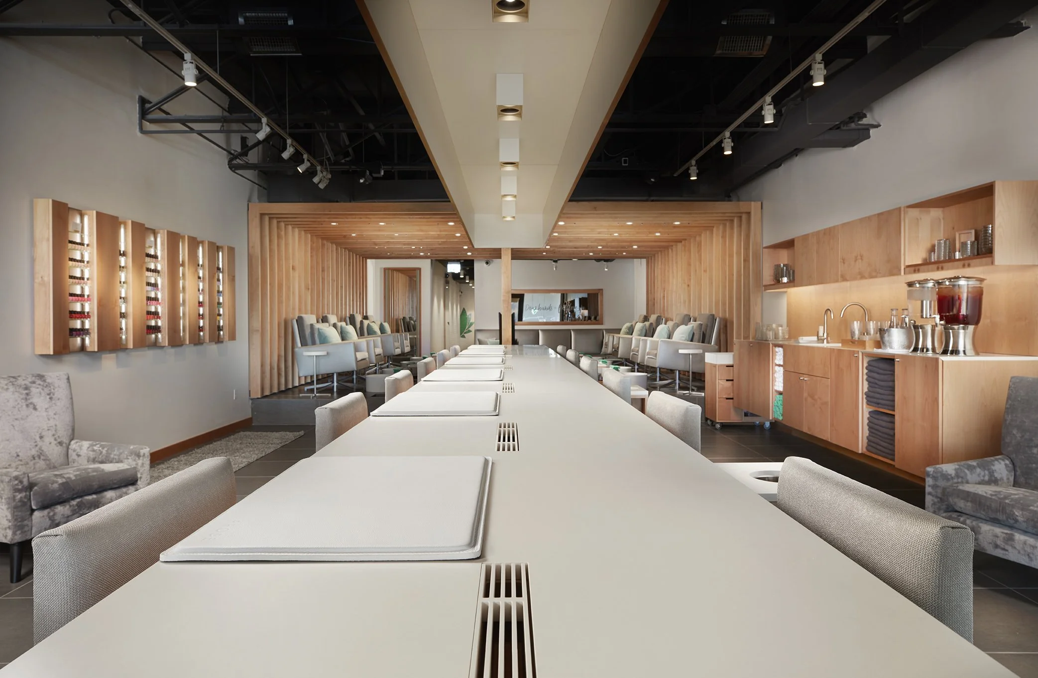 Modern interior of a spa or lounge with a long white table, chairs, cushioned chairs along the walls, wooden accents, and a beverage station.