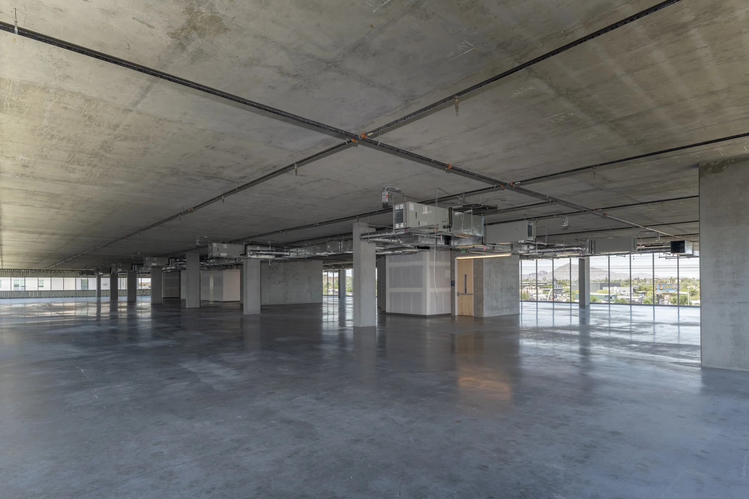 Empty commercial building with concrete floors, unfinished walls, and exposed HVAC and piping system on the ceiling. Large windows show a cityscape outside.