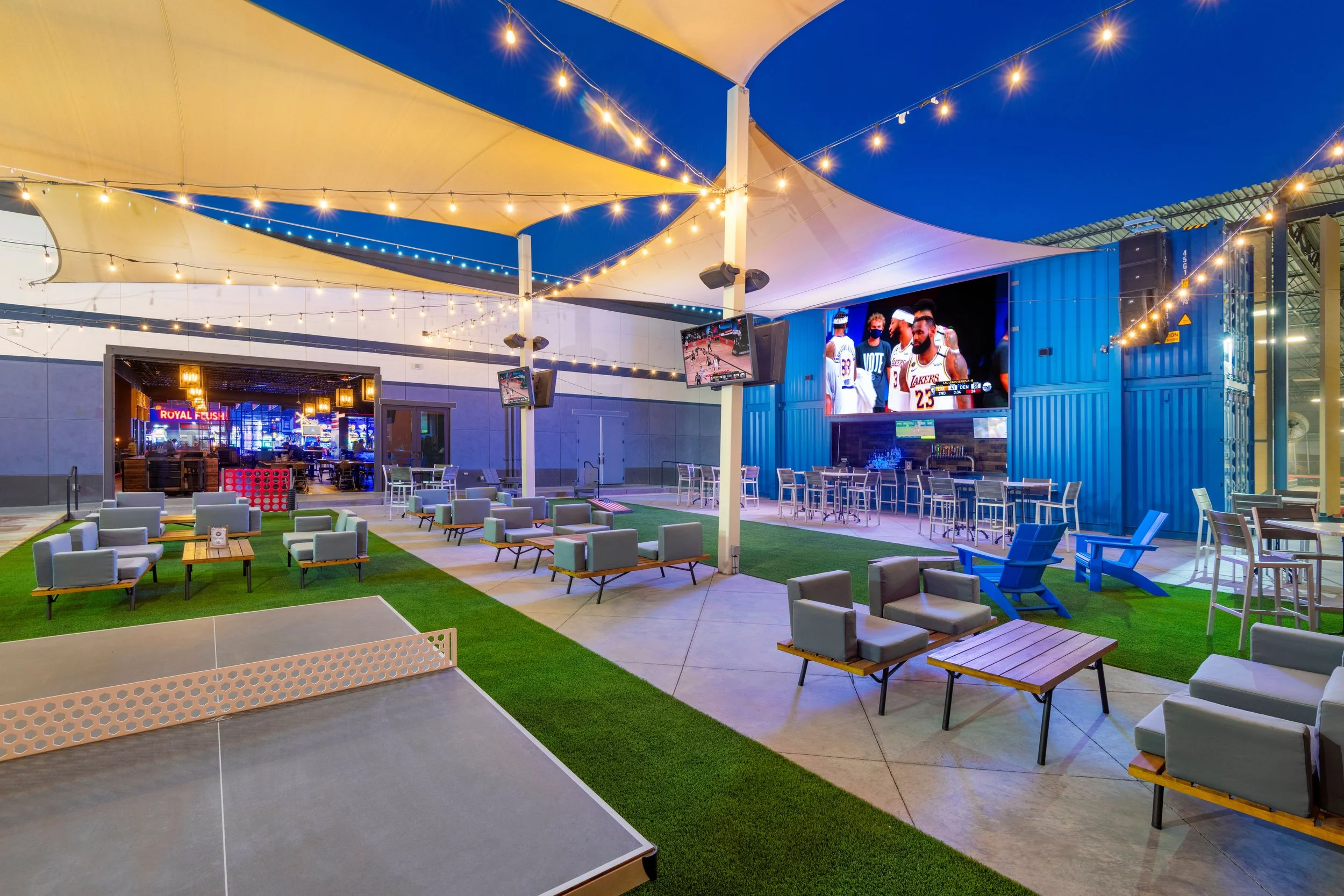 An outdoor lounge area with string lights, multiple TV screens showing a basketball game, and seating arrangements including chairs and tables. There's a ping pong table in the foreground.