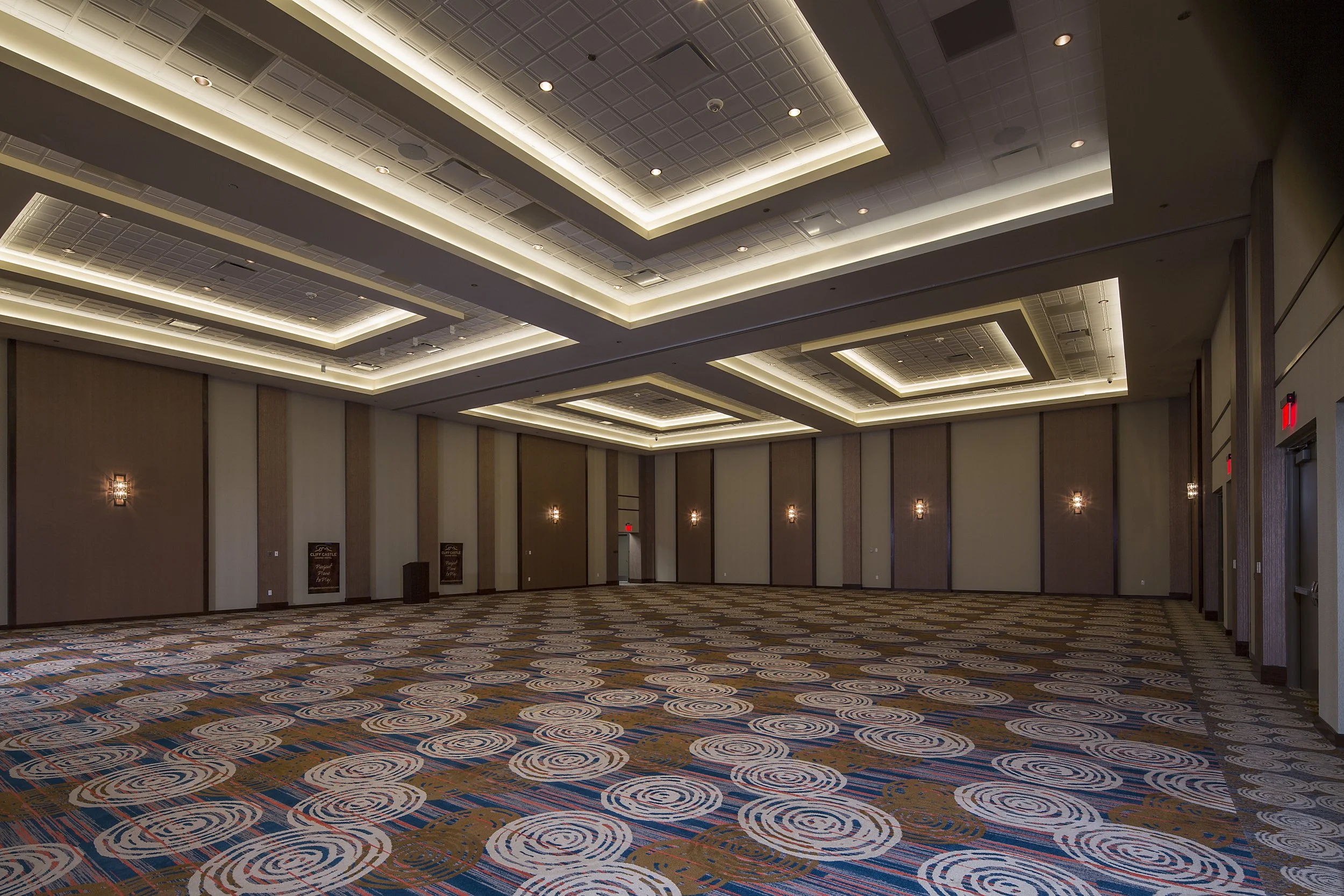 Empty event ballroom with patterned carpet, beige and brown walls, wall sconces, and recessed lighting in the ceiling.