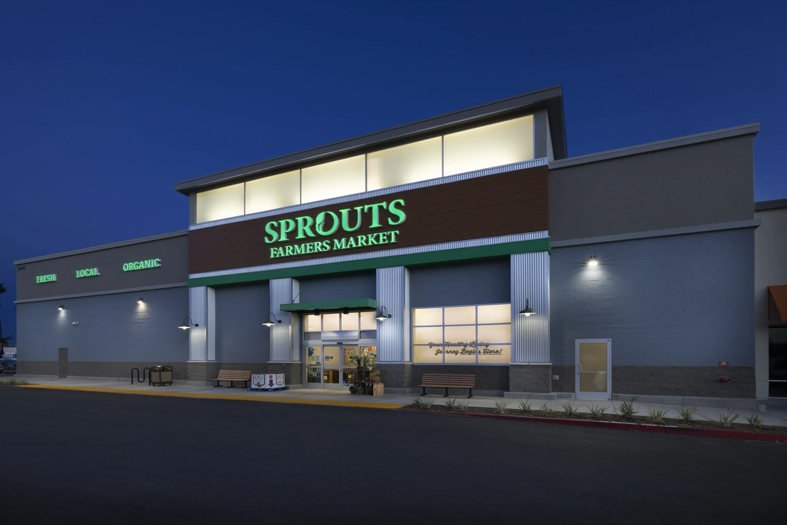 Exterior of Sprouts Farmers Market grocery store at dusk, with green illuminated signage and a parking lot in the foreground.