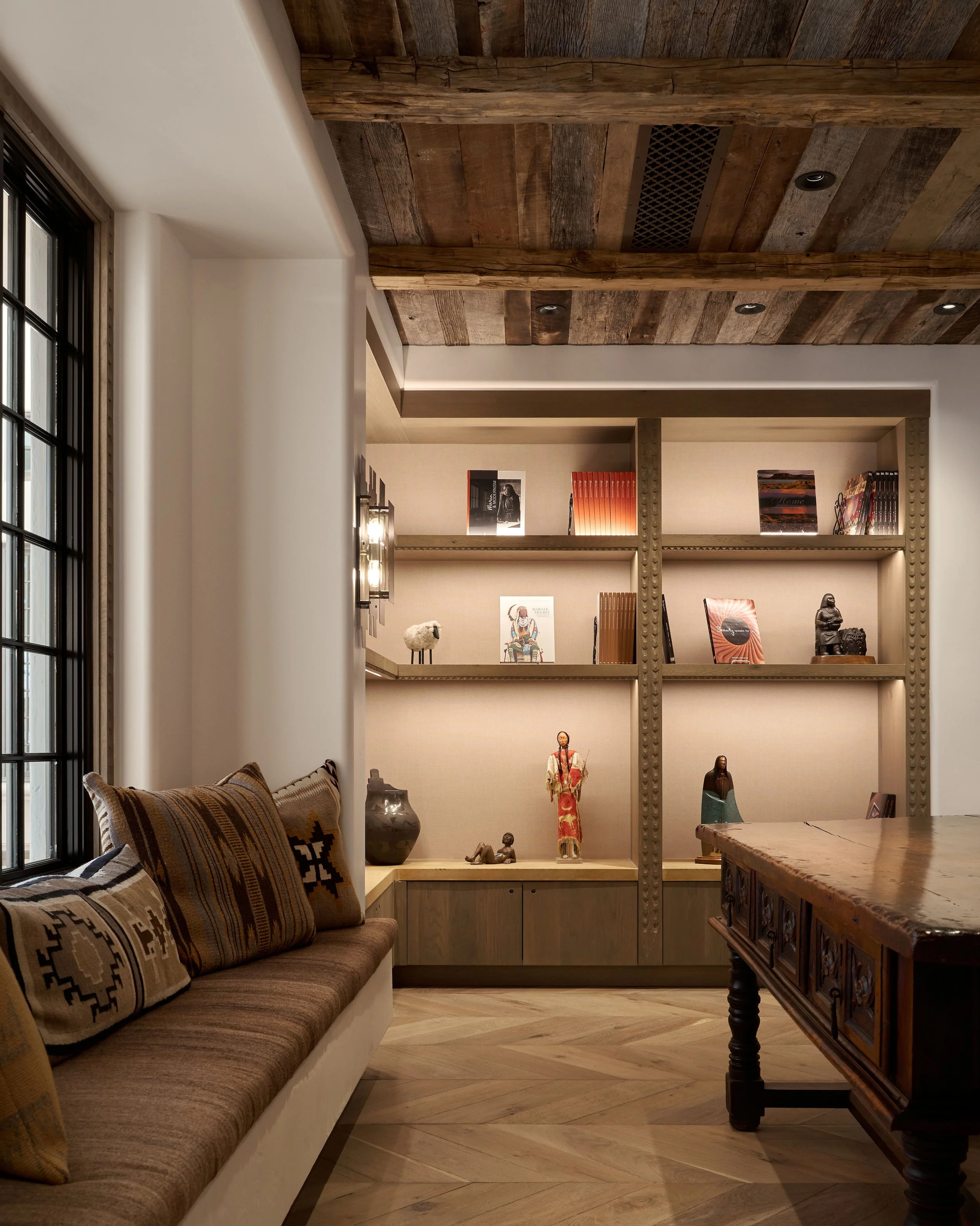 Cozy living room with a built-in bookshelf displaying books and decorative items, a wooden ceiling, a window with black frame, and a sofa with patterned pillows.