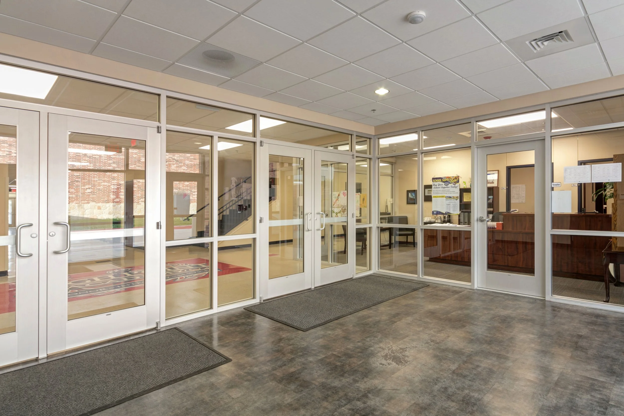 Indoor view of a school entrance with glass double doors and a reception area visible through glass walls.