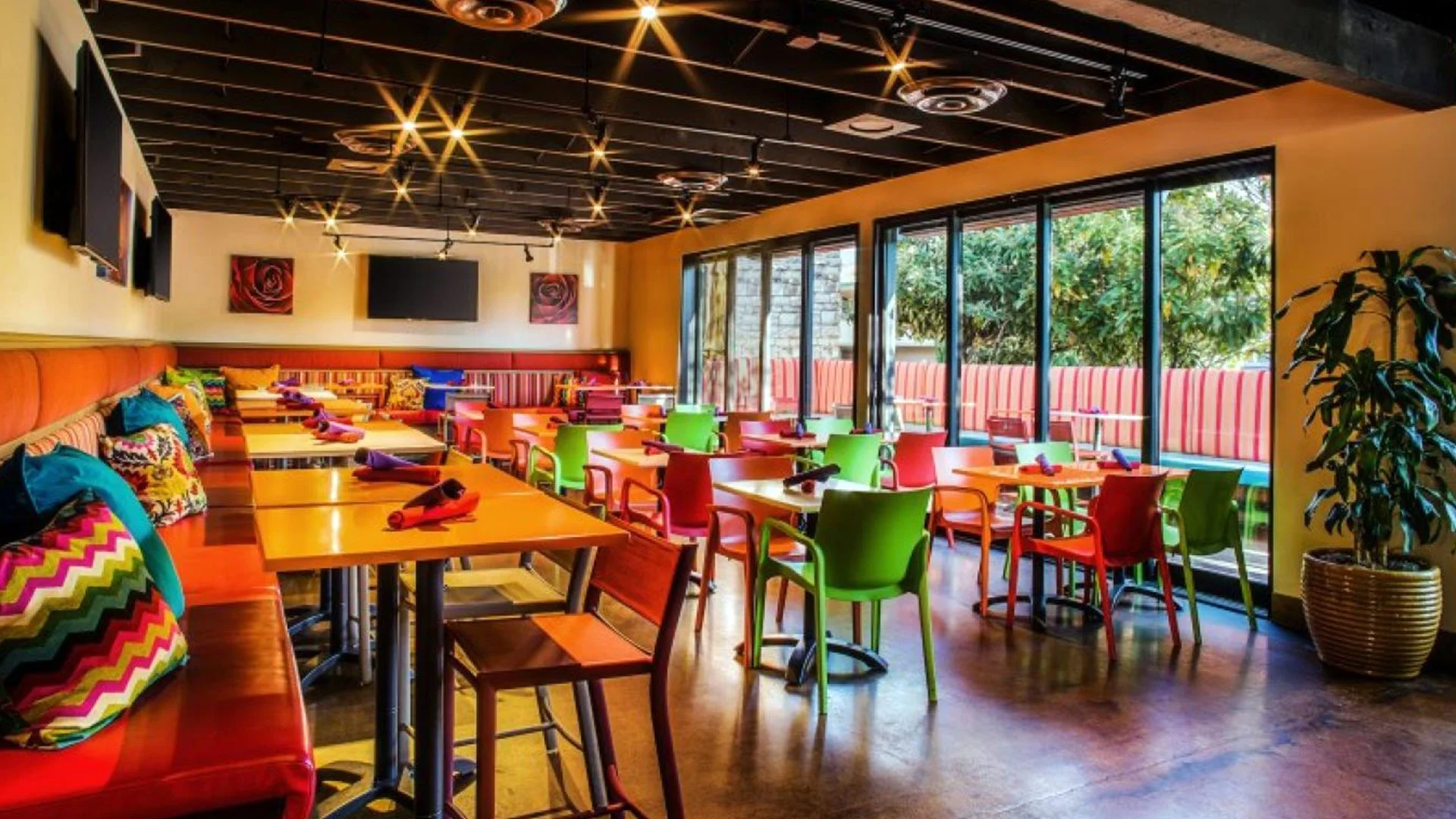 Colorful dining area with vibrant chairs and cushions, large windows with outdoor greenery, and ceiling lights.