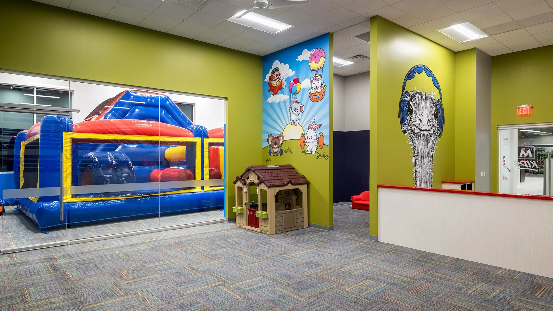 Indoor play area with a colorful inflatable bounce house, a small playhouse, and vibrant wall murals of animals and cartoon characters, in a family-friendly entertainment center.