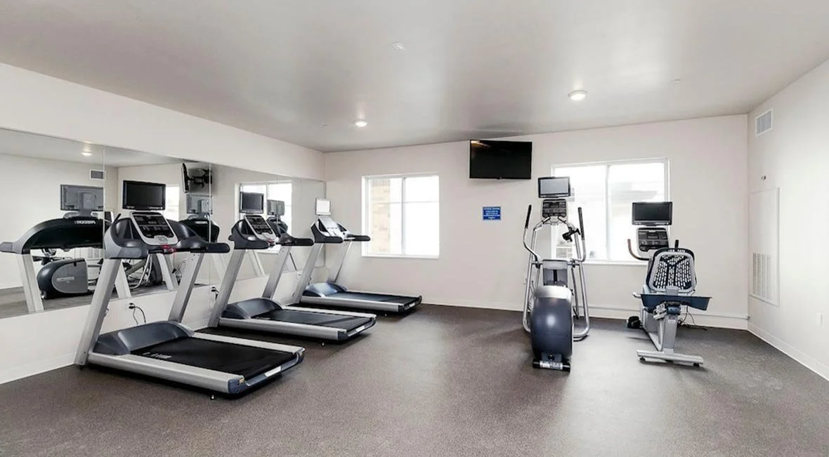 Empty gym room with cardio machines including treadmills, stationary bikes, and elliptical trainers, facing a mirror and two windows.