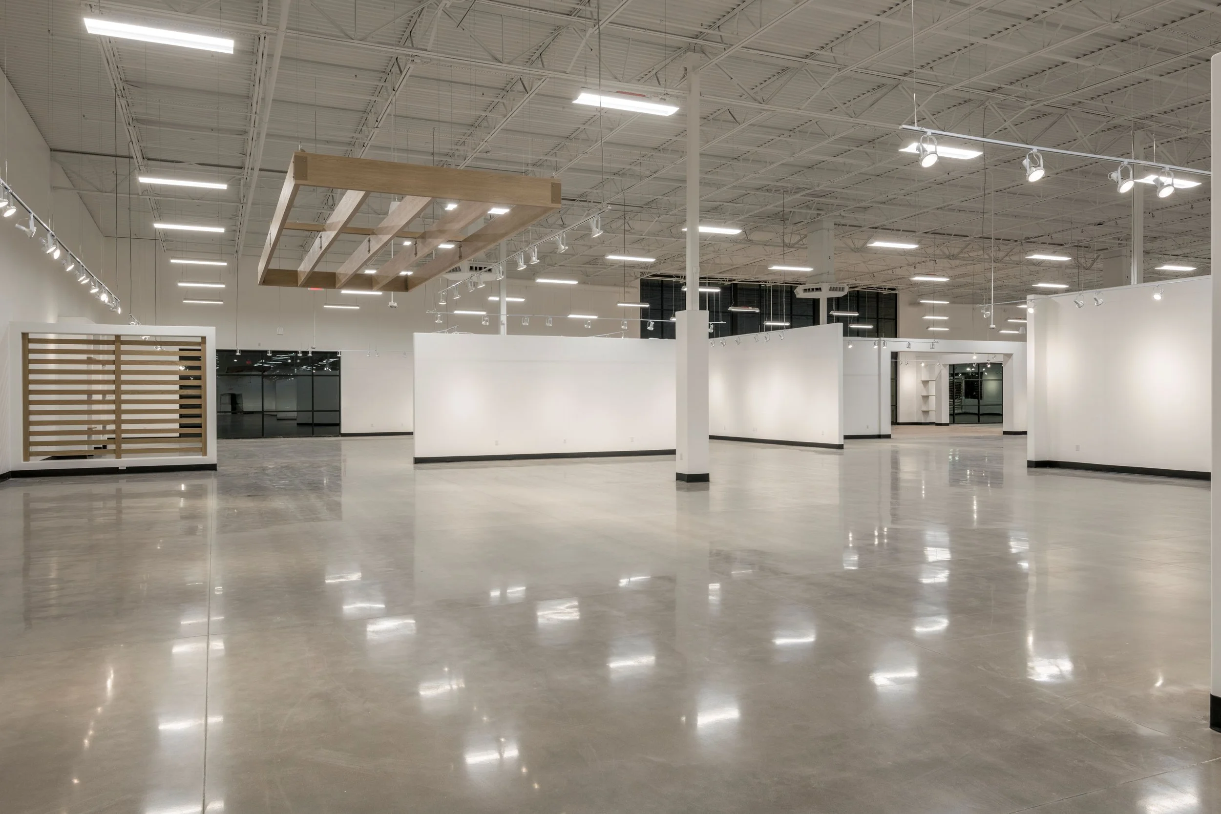 Empty retail or gallery space with polished concrete floors, white walls, and ceiling with exposed ductwork and lighting fixtures. There are some partitions and a wooden structure hanging from the ceiling.