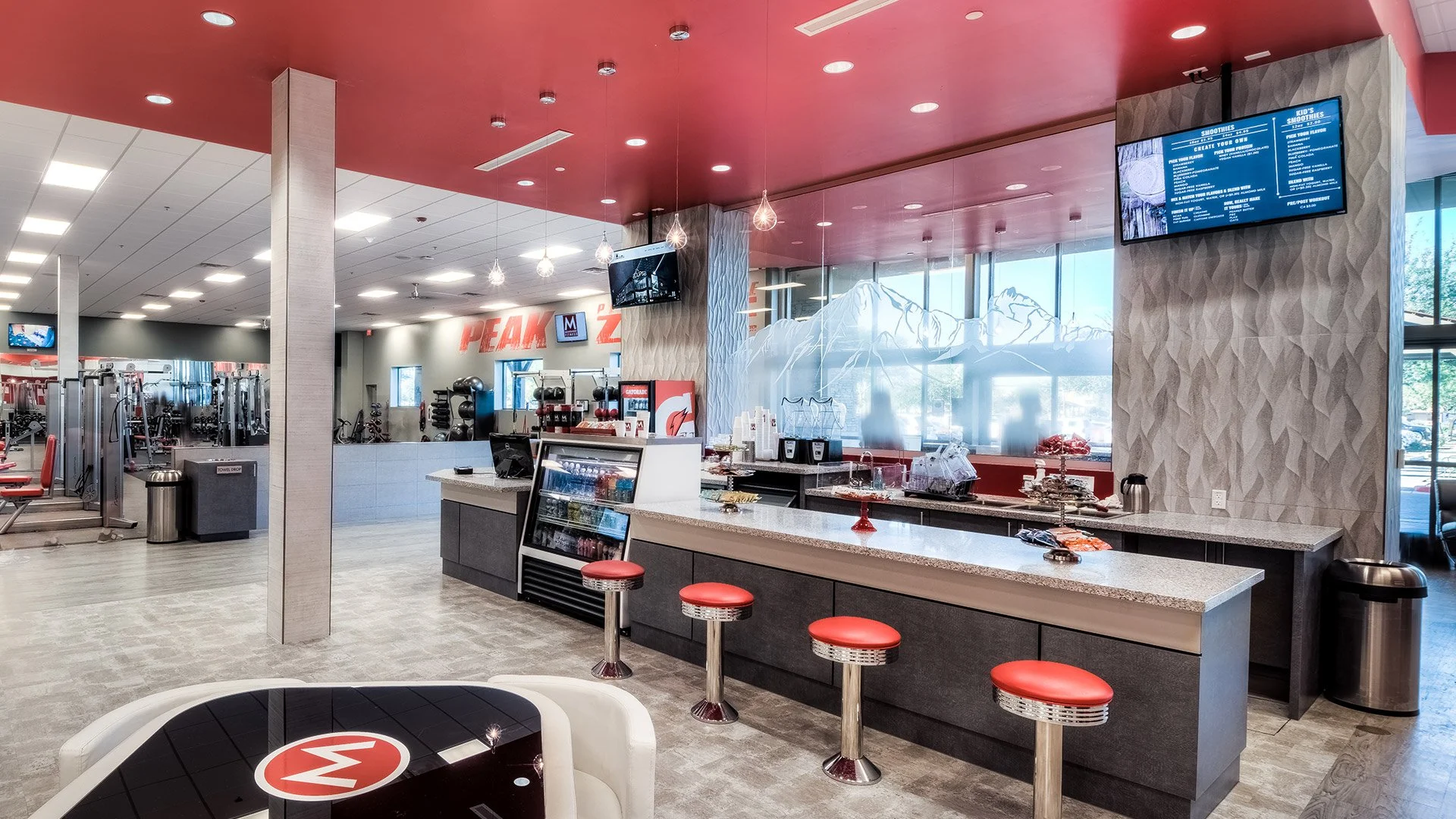 A modern gym with a refreshment bar and seating area near windows, and gym equipment visible in the background.