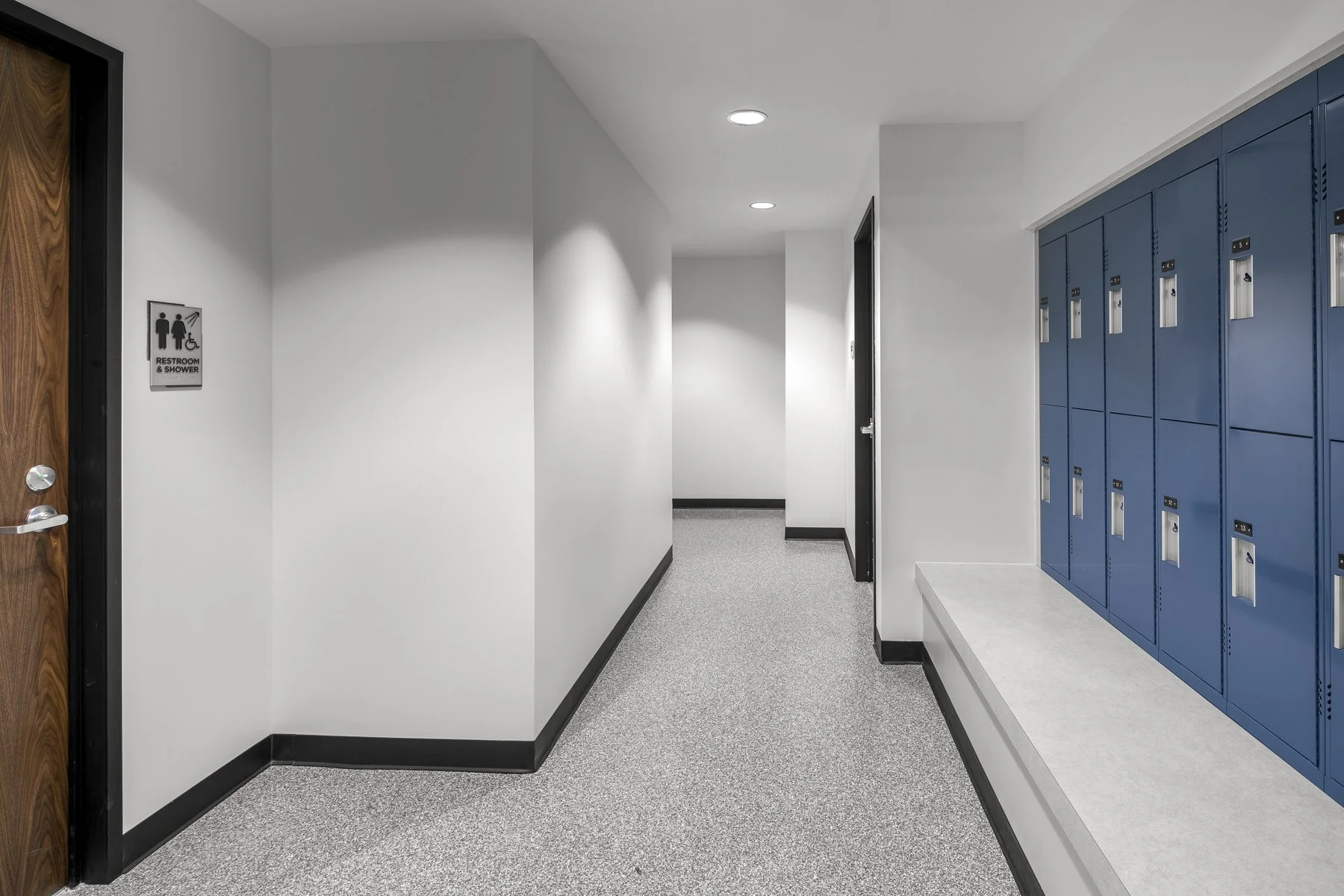 School hallway with lockers on the right, a door with a restroom sign on the left, and a white wall with ceiling lights.
