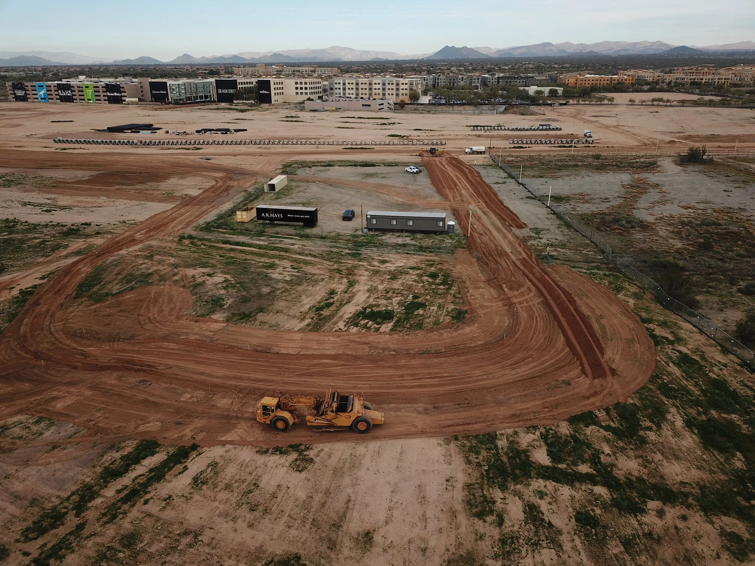 An aerial view of a construction site with a large dirt area, a bulldozer, and a temporary building, with an urban area and mountains in the background.