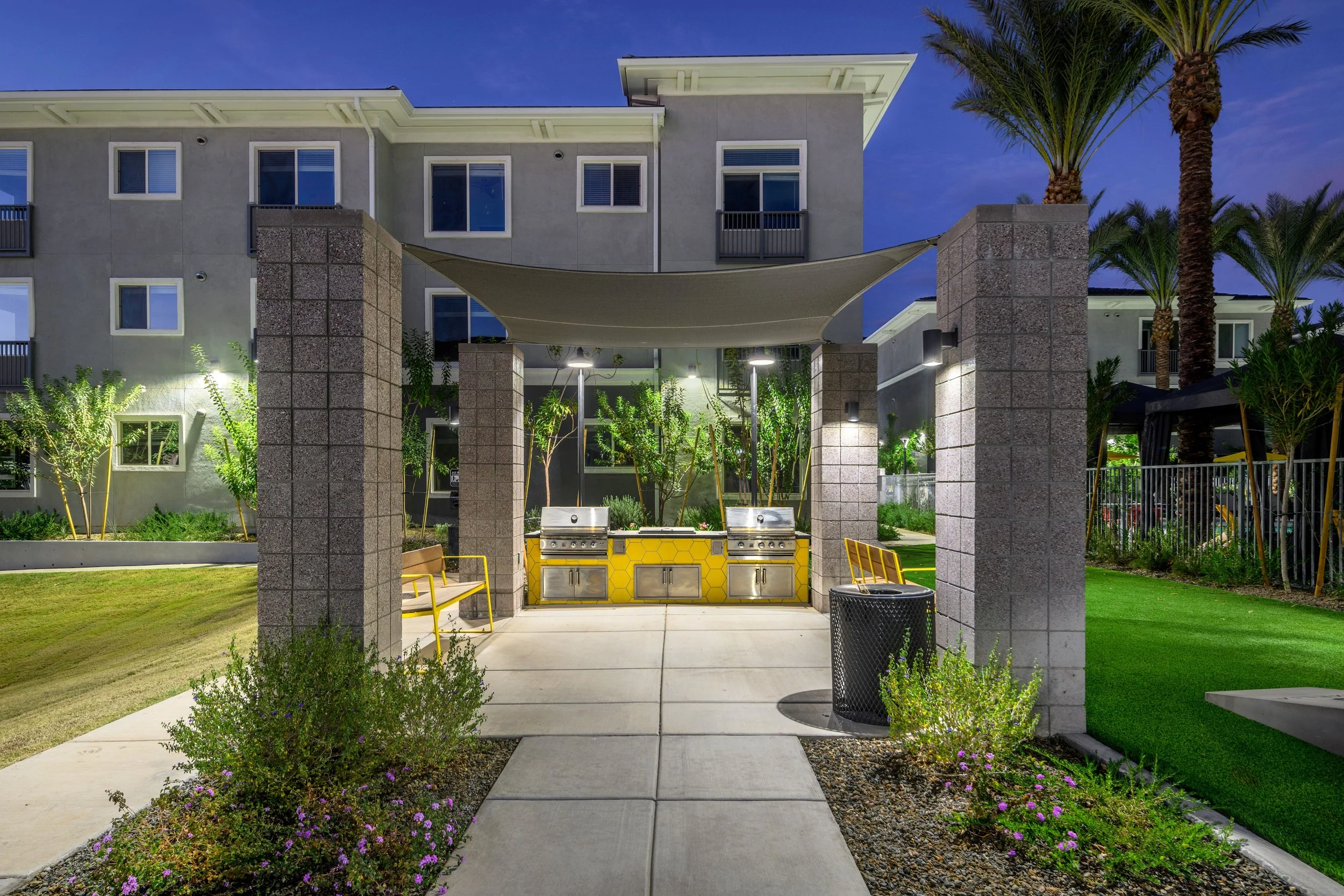 Outdoor community barbecue area with built-in grills, yellow honeycomb-patterned cabinets, surrounded by palm trees and greenery at dusk.