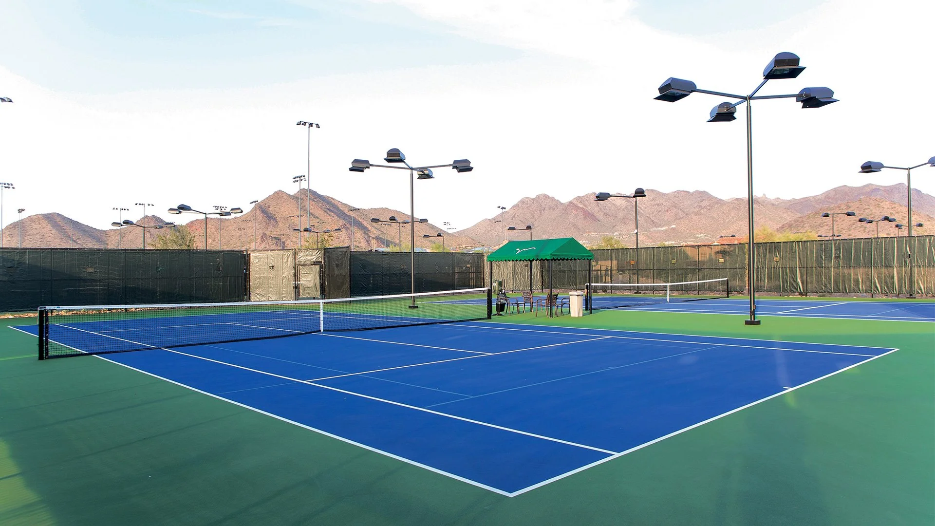Empty outdoor tennis courts with blue playing surfaces, surrounded by black wind screens, mountain range in the background, and multiple tall lamps overhead. A small shaded area with chairs and a trash can is near the courts.