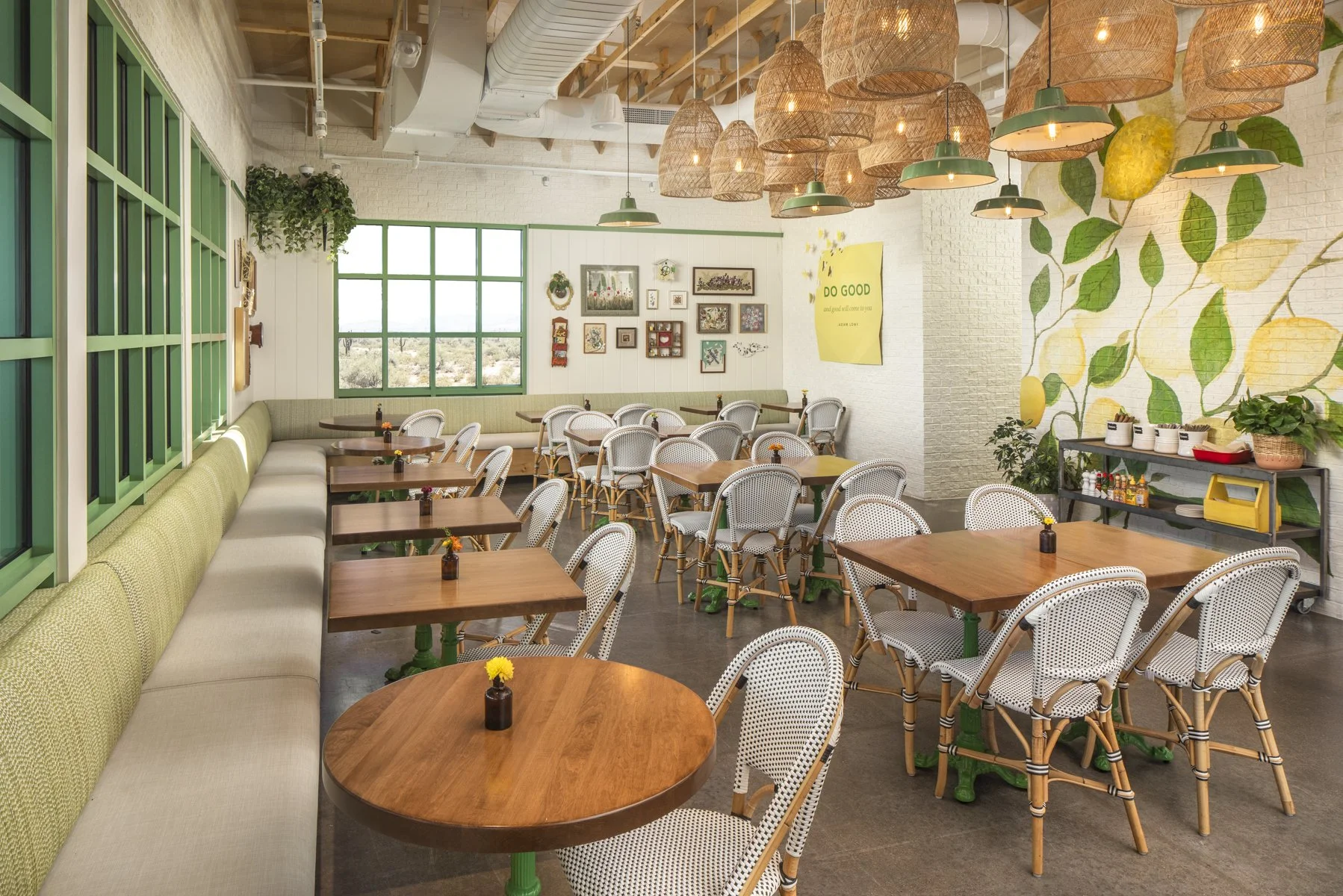 Cozy cafe with green window frames, white brick walls, an assortment of framed art, a large wall mural of lemon and leaves, and hanging wicker pendant lights. Wooden tables and white wicker chairs are arranged throughout, some with small flower vases