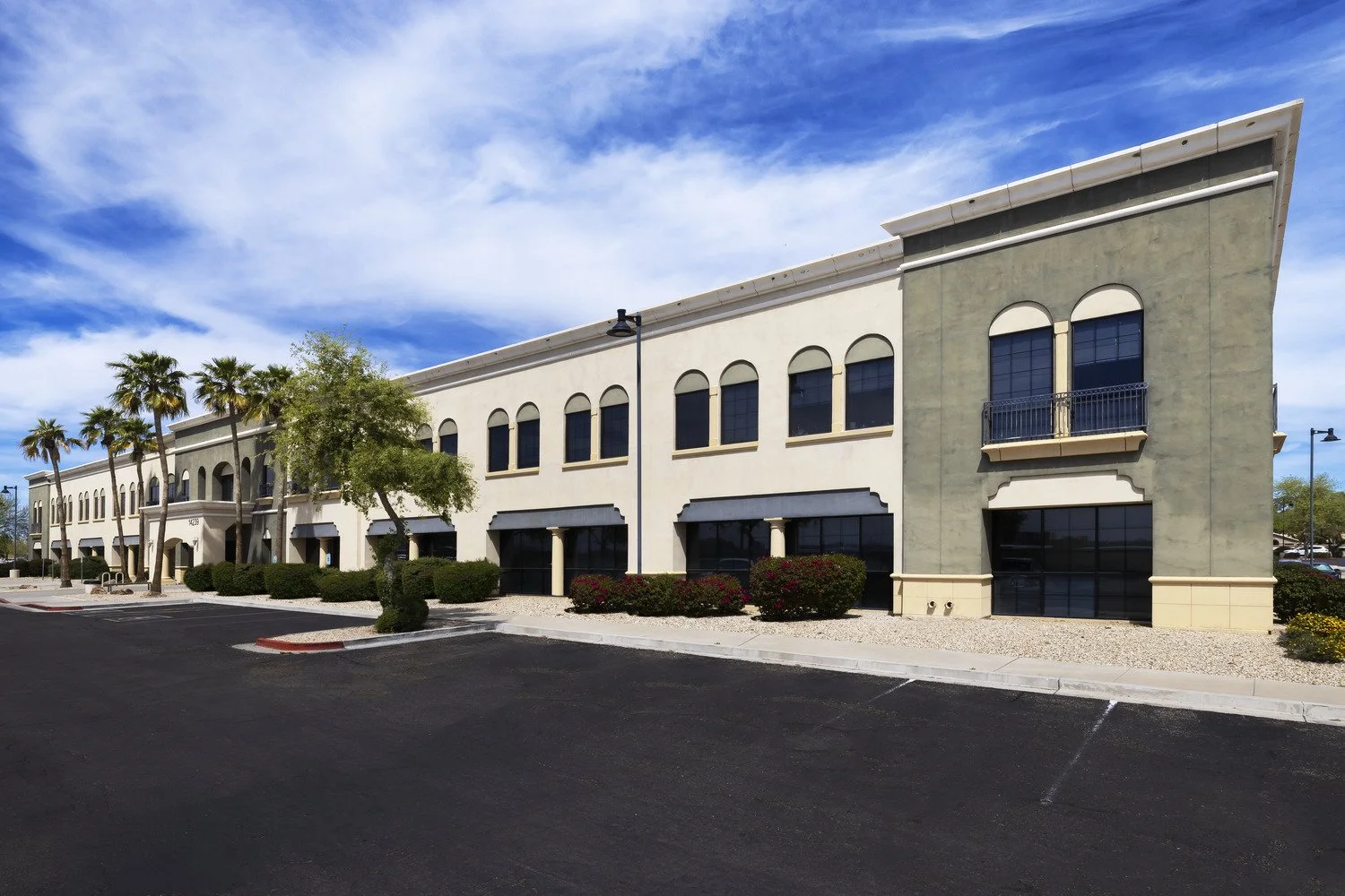 A commercial shopping center with a beige and light gray facade, black storefronts, and a few small trees and bushes in the parking lot. The sky is partly cloudy with blue and white clouds.