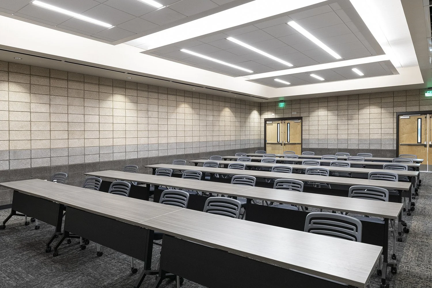 Empty classroom or conference room with long tables and gray chairs, beige brick walls, and double wooden doors, illuminated by ceiling lights.
