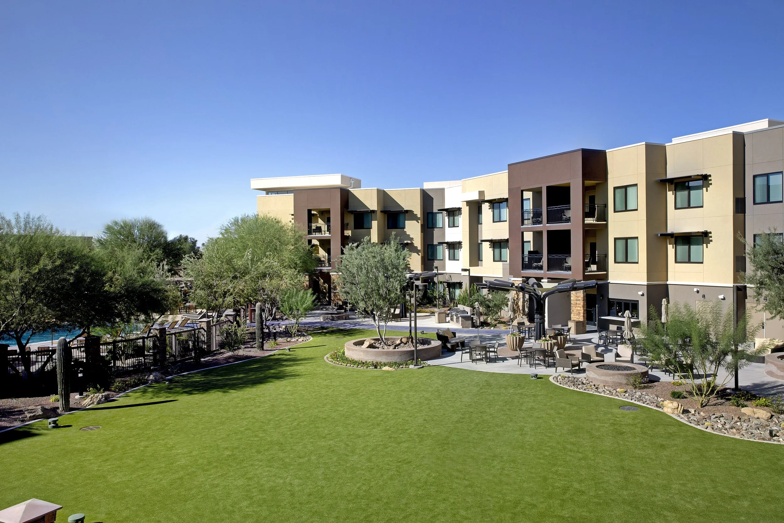 Apartment complex with landscaped courtyard featuring green grass, trees, outdoor seating, and a swimming pool under a clear blue sky.