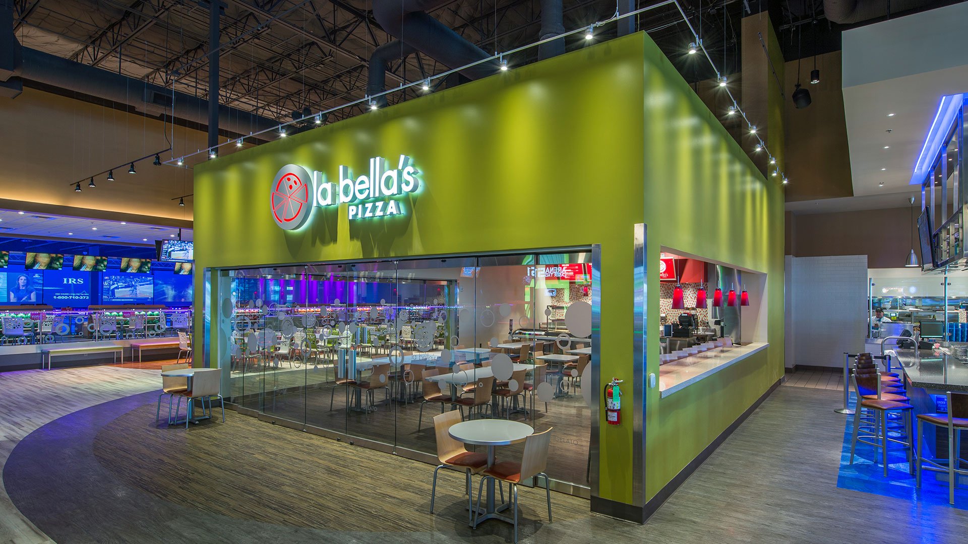 Inside La Bella's Pizza restaurant with green exterior walls, glass windows, seating area, and colorful lighting.