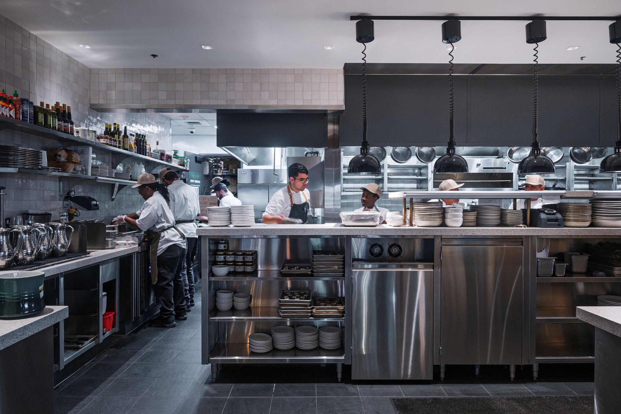 Commercial kitchen with chefs preparing dishes behind a counter, black pendant lights, and stainless steel appliances.