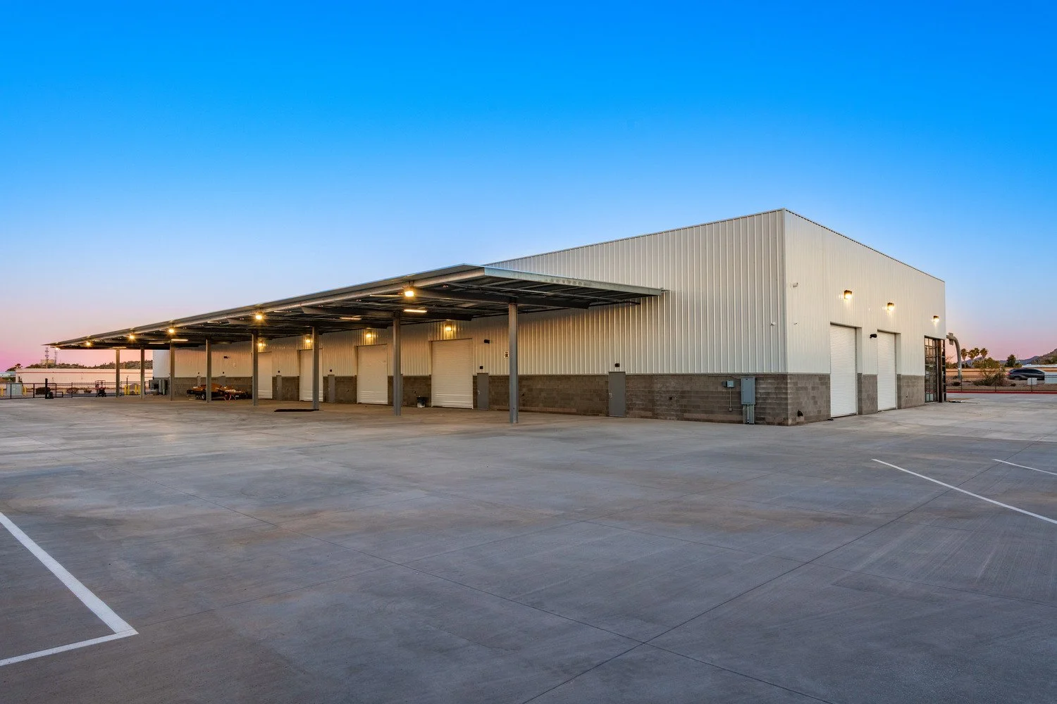 A large empty parking lot in front of a warehouse-style building at sunset. The building has a white metal exterior with a row of overhead doors and exterior lighting.