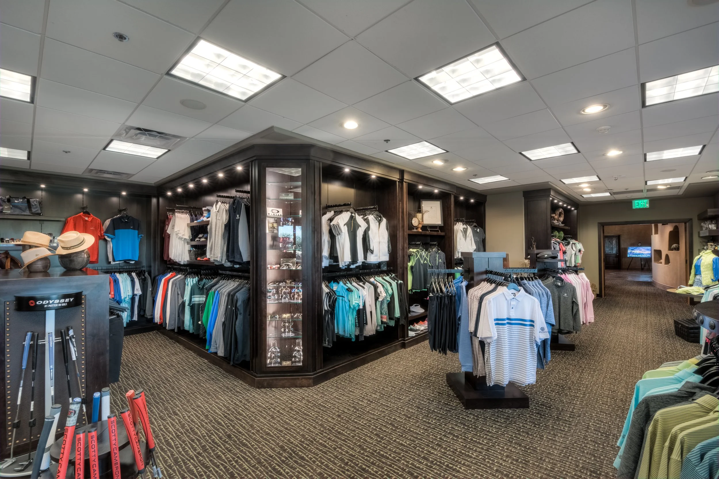 Interior view of a golf apparel store with clothing racks and displays of golf shirts, hats, and golf accessories, illuminated by overhead lighting and carpeted flooring.