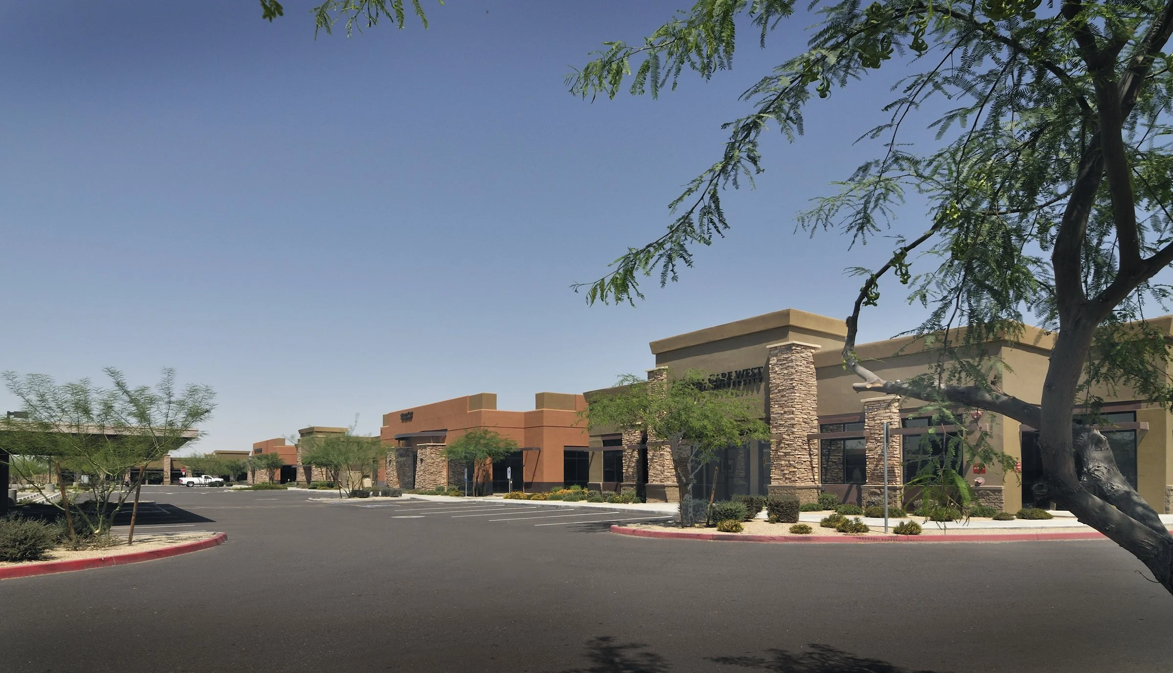 Empty shopping center parking lot with trees, storefronts, and a clear blue sky.