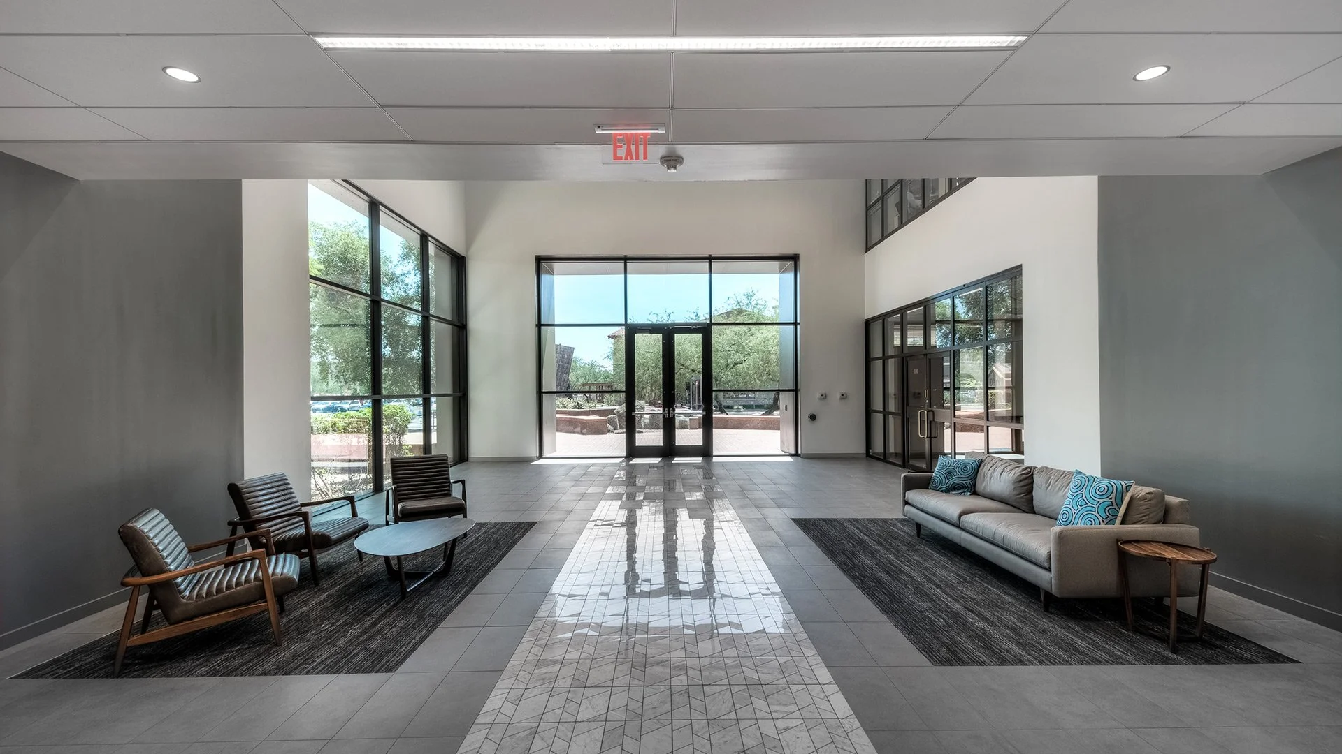Modern lobby area with large glass windows, a beige sofa with blue patterned pillows, a small side table, black chairs with striped cushions, tiled floor, and an exit door with an illuminated exit sign above.