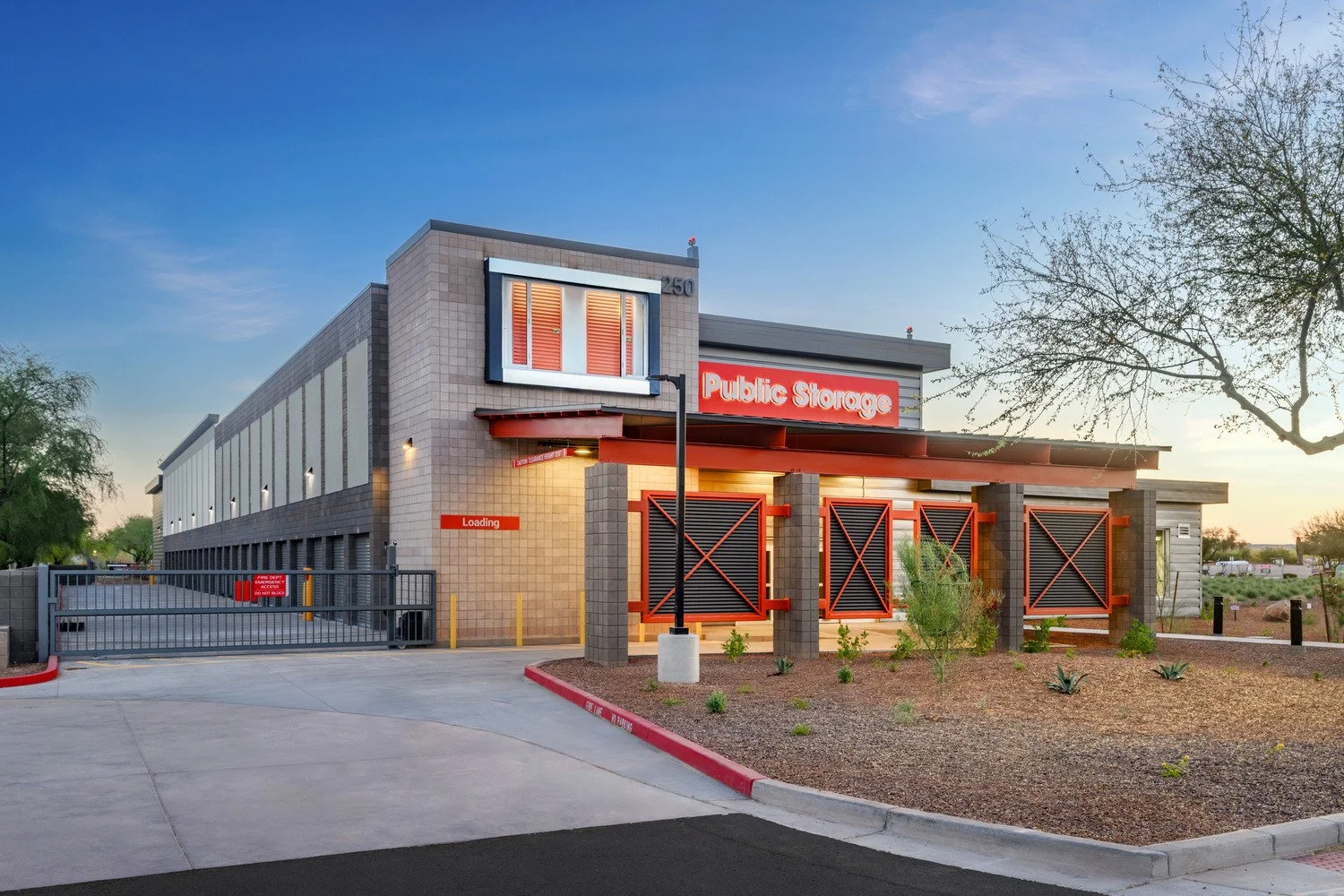 Exterior of a self-storage facility with a sign reading 'Public Storage,' a loading area, and a gate, during early evening with a clear sky and desert landscape.