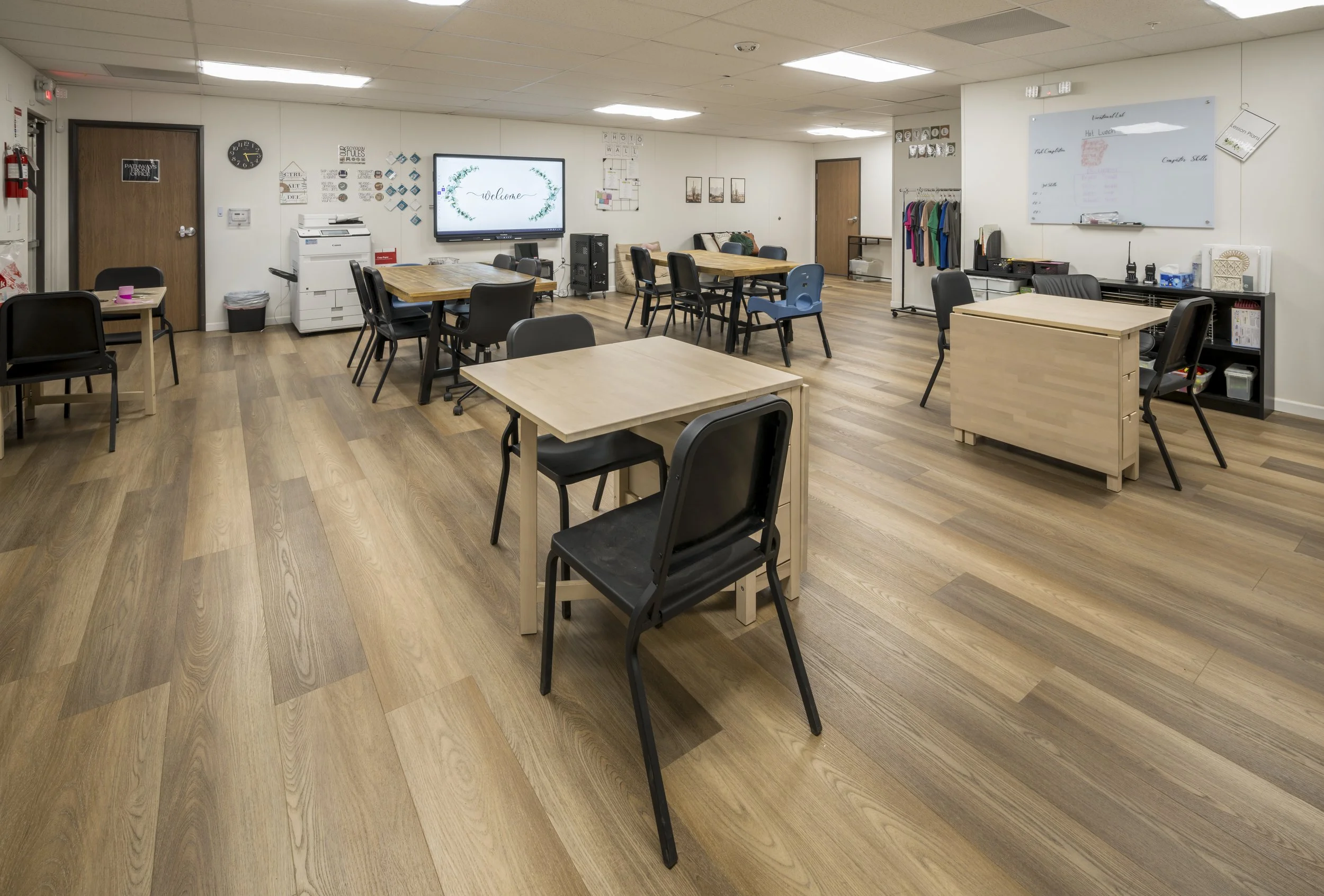 Empty classroom or meeting room with tables, chairs, a whiteboard, and a large screen display on a wall.