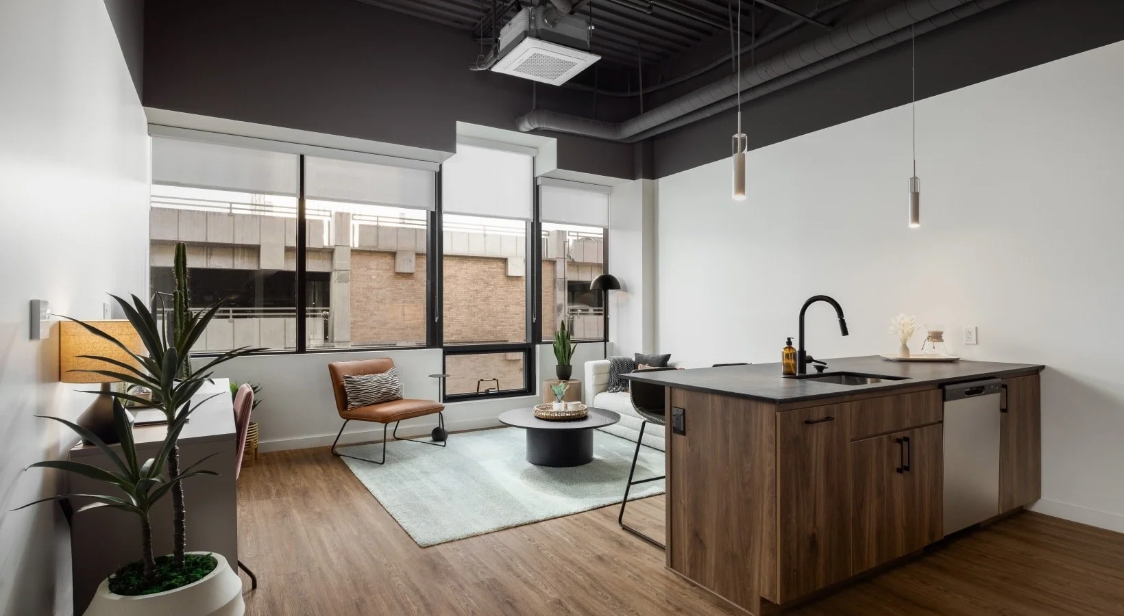Open-concept living room and kitchen with large windows, seating area with chairs and a rug, and a kitchen island with a sink and pendant lights.