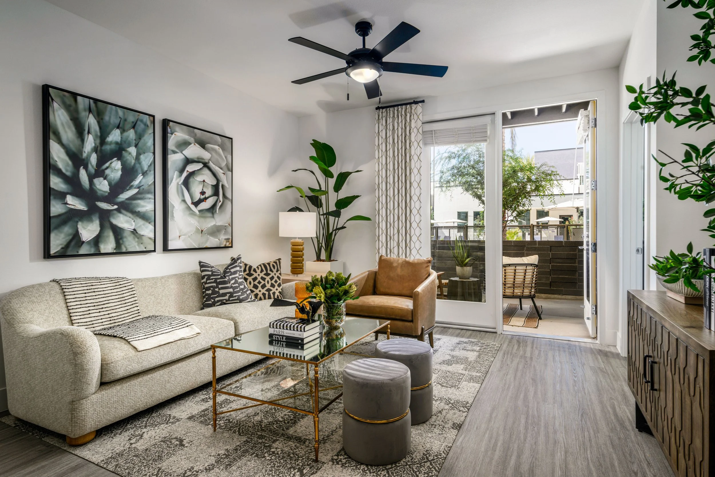 Living room with beige sofa, leather armchair, glass coffee table, patterned rug, large potted plants, wall art of succulents, window with curtains, balcony outside, and gray wood flooring.
