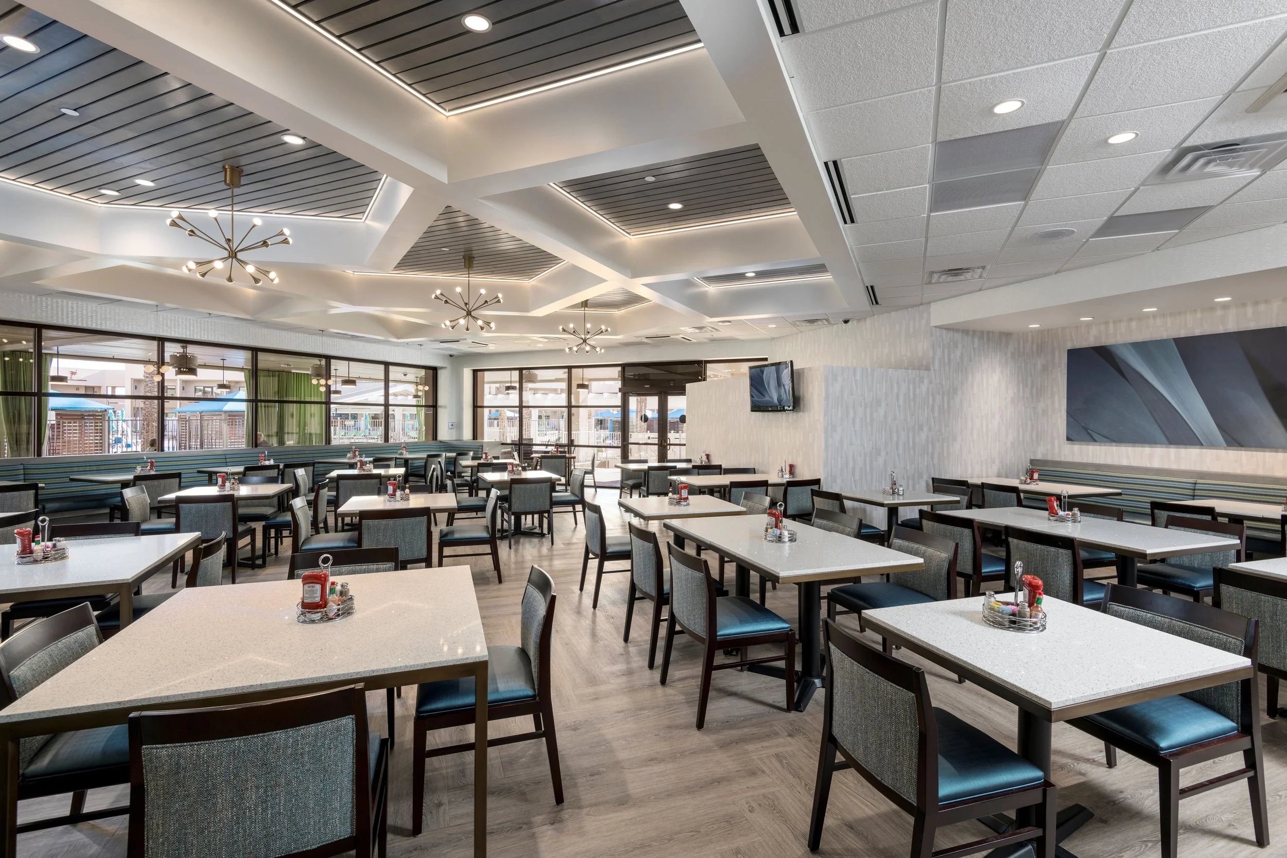 Empty restaurant dining area with modern lighting, tables with condiments, and large windows.