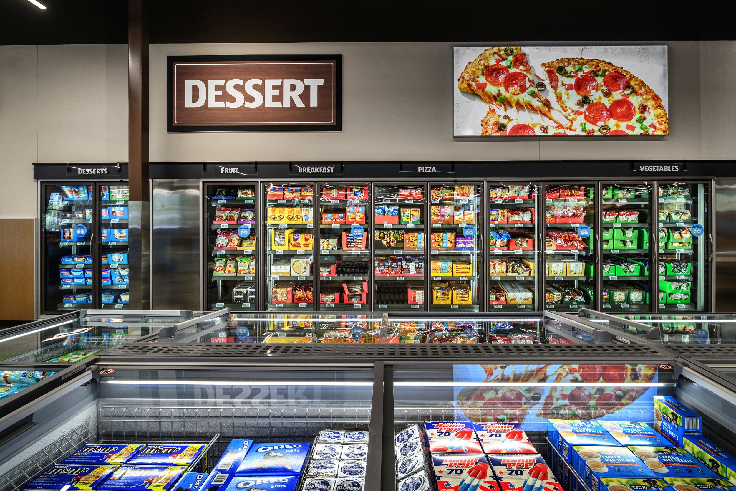 Refrigerated snack aisle in a grocery store with sections for desserts, fruit, breakfast, pizza, and vegetables, and a sign overhead labeled 'DESSERT' above a digital display of pizza.