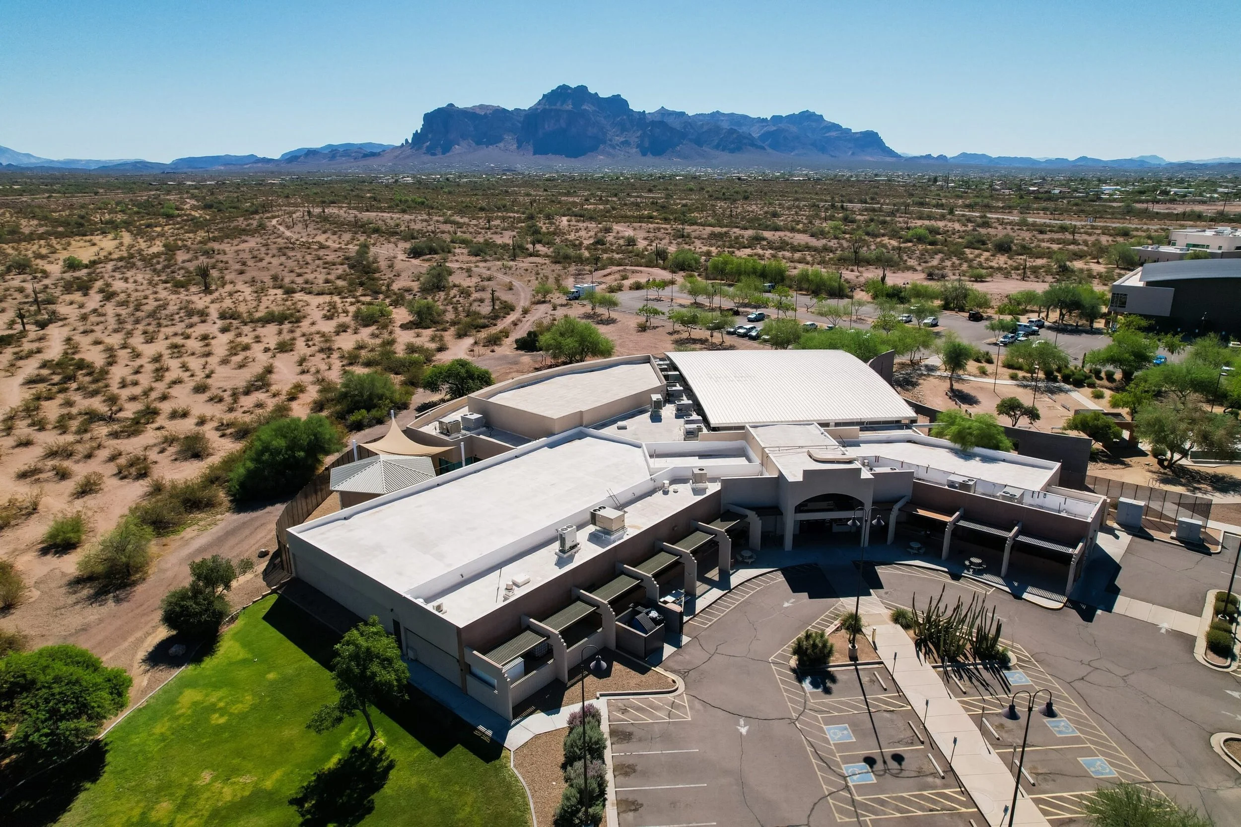 Aerial view of a large modern building with a parking lot surrounded by desert landscape and mountains in the background.