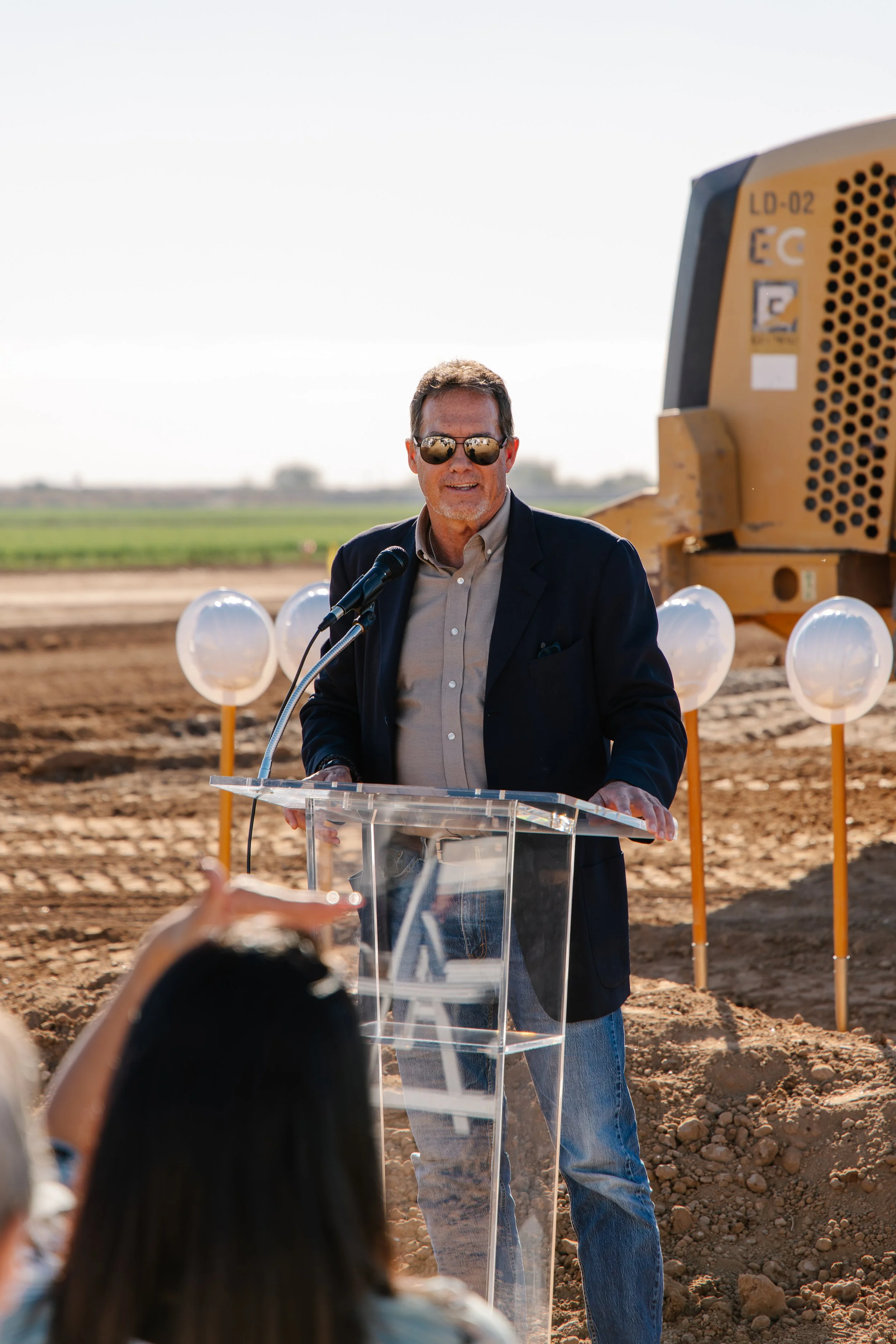 Adam Mays in a suit jacket, sunglasses, and jeans speaking at a podium during a groundbreaking ceremony at a construction site with earth-moving equipment and hard hats and shovels in the background.