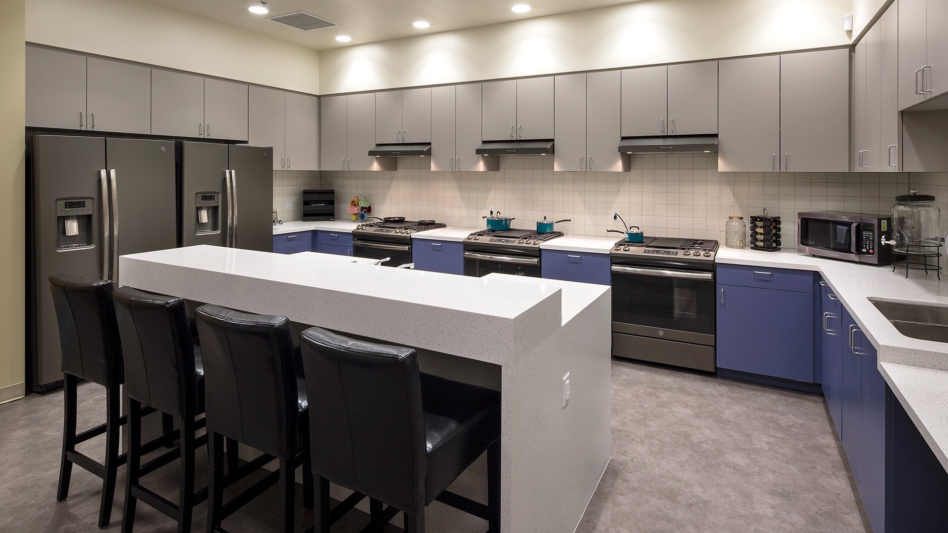 Kitchen with gray and blue cabinets, stainless steel refrigerator, white countertop island, black chairs, and various small appliances like microwave, pots, and spice rack.