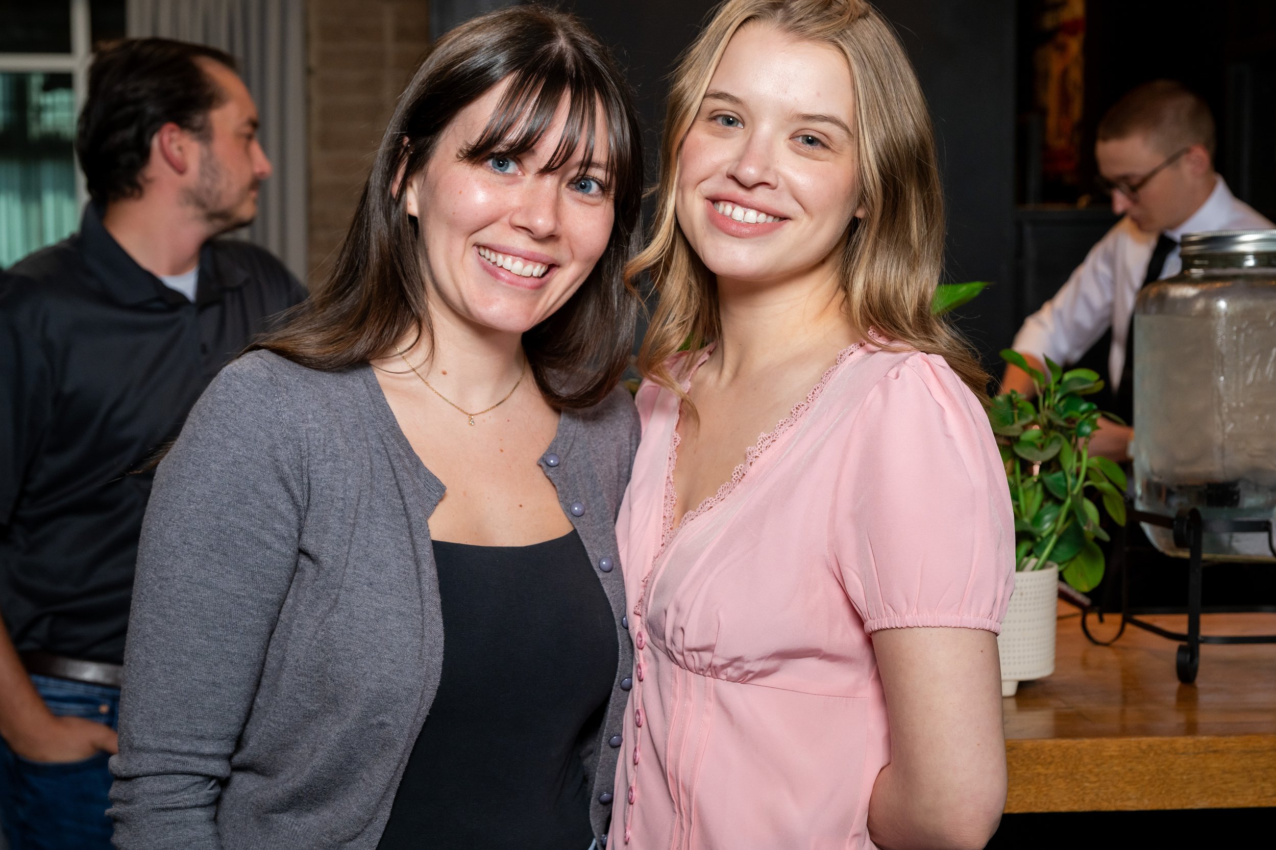 Two women smiling at the camera at a social gathering, with a man in the background and a bar area behind them.