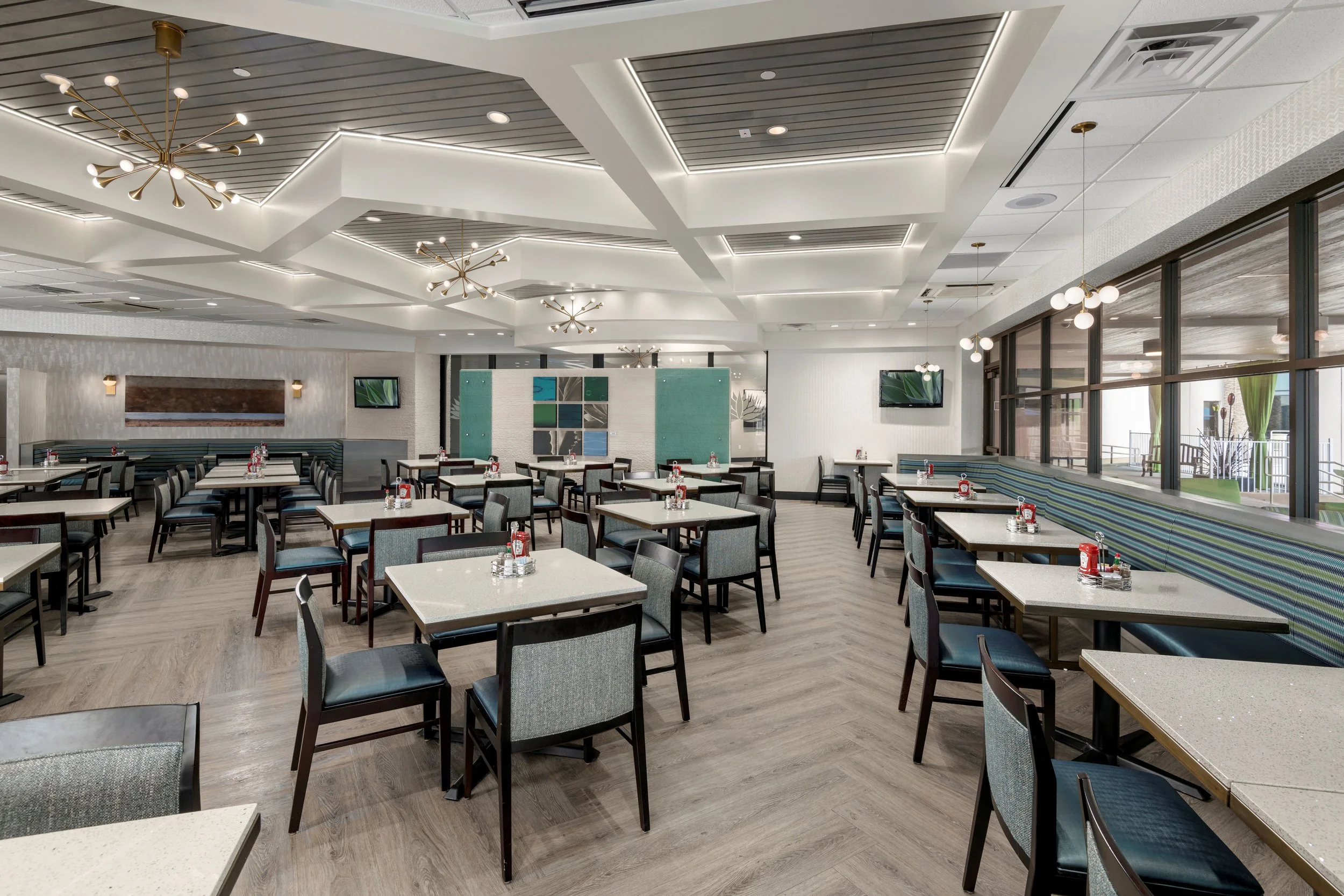 Empty dining area with tables and chairs, modern lighting fixtures, large windows, and a TV on the wall.
