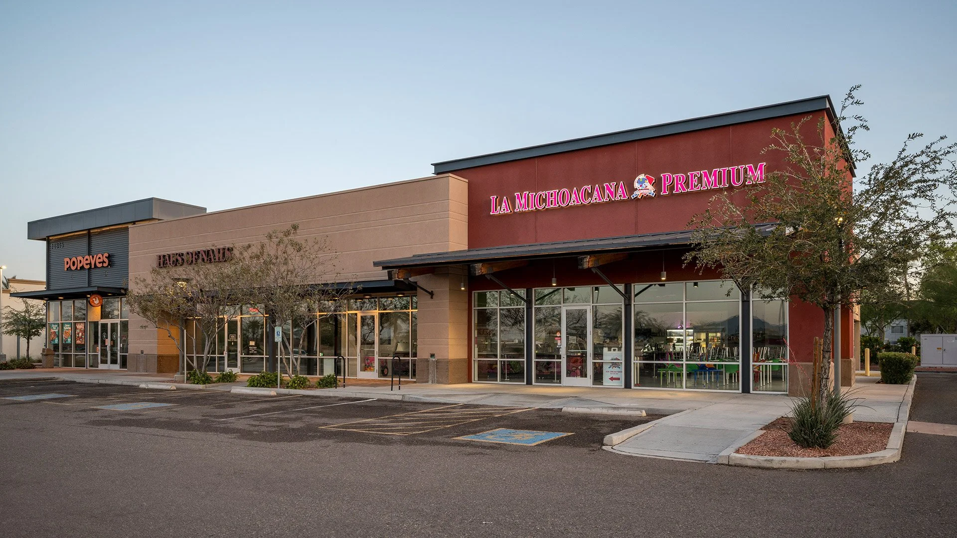 A shopping plaza with La Michoacana Premium, Popeyes, and Has O'Fails storefronts, parking lot, trees, and a clear sky.