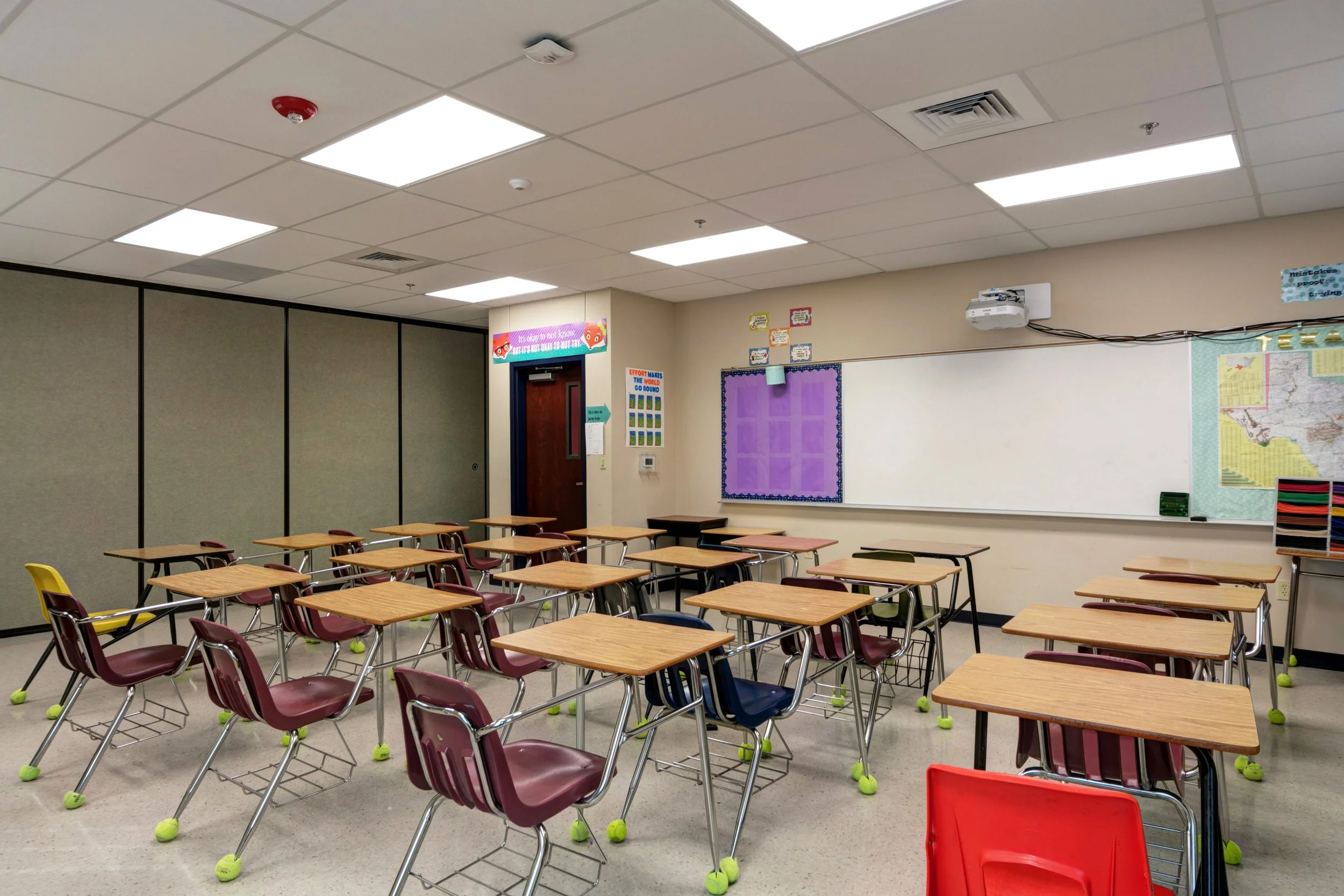 Empty classroom with student desks, chairs, a whiteboard, educational posters, and a projector on the ceiling.