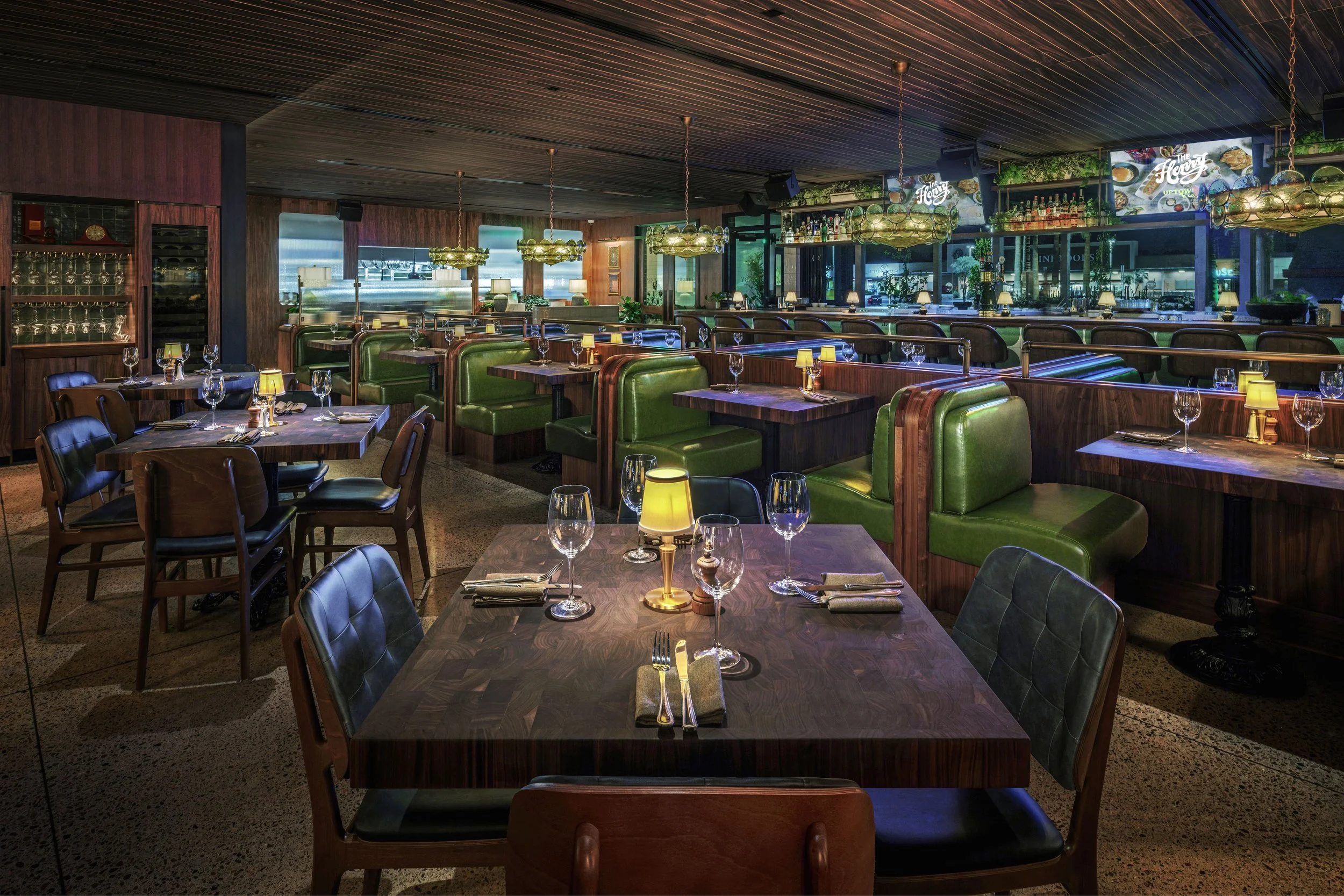 Interior of a modern restaurant with wooden tables, green leather booth seating, glassware, and small lamps on the tables. The bar area is visible in the background with bottles and bar signage.